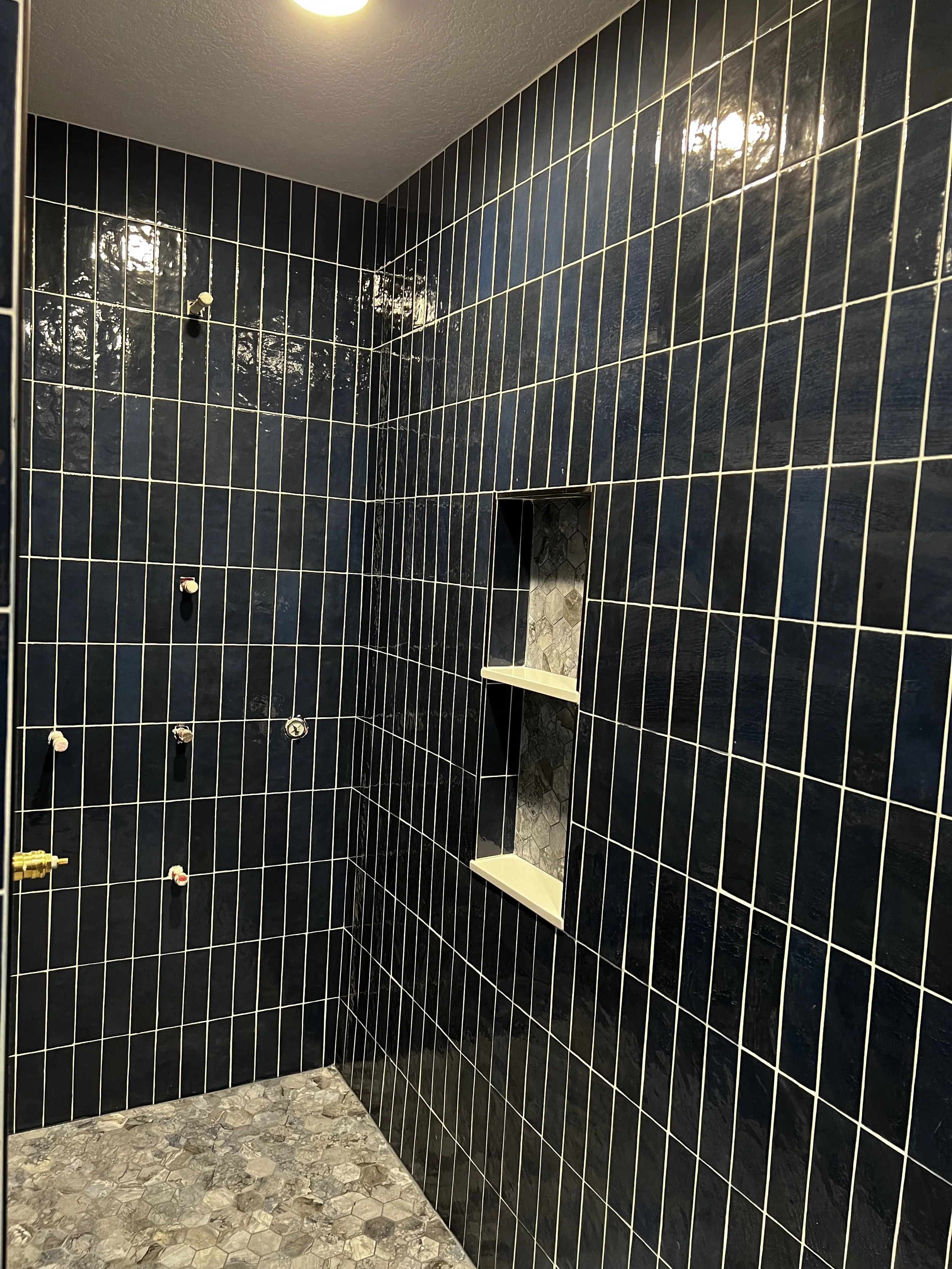 A bathroom shower with black tiles, a built-in soap shelf, and plumbing fixtures on the wall, with a yellow ceiling light.
