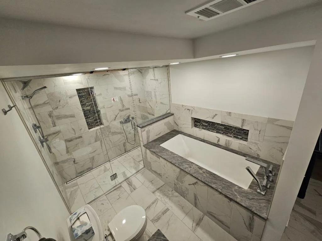Modern bathroom with marble tiles, glass shower enclosure with multiple shower heads, a bathtub with a granite surround, and a toilet, all in neutral tones.