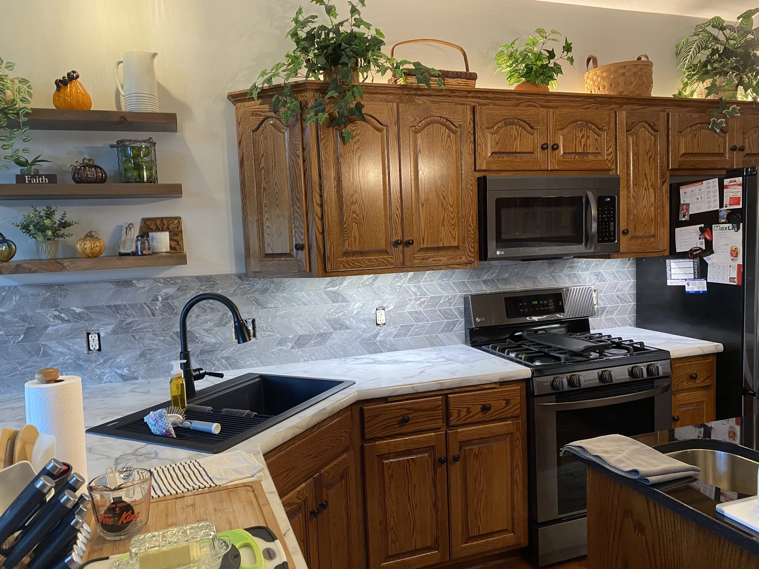 Kitchen with wooden cabinets, marble countertop, stainless steel oven and microwave, black sink with a black faucet, and various kitchen items and decorations, including plants and shelving on the wall.