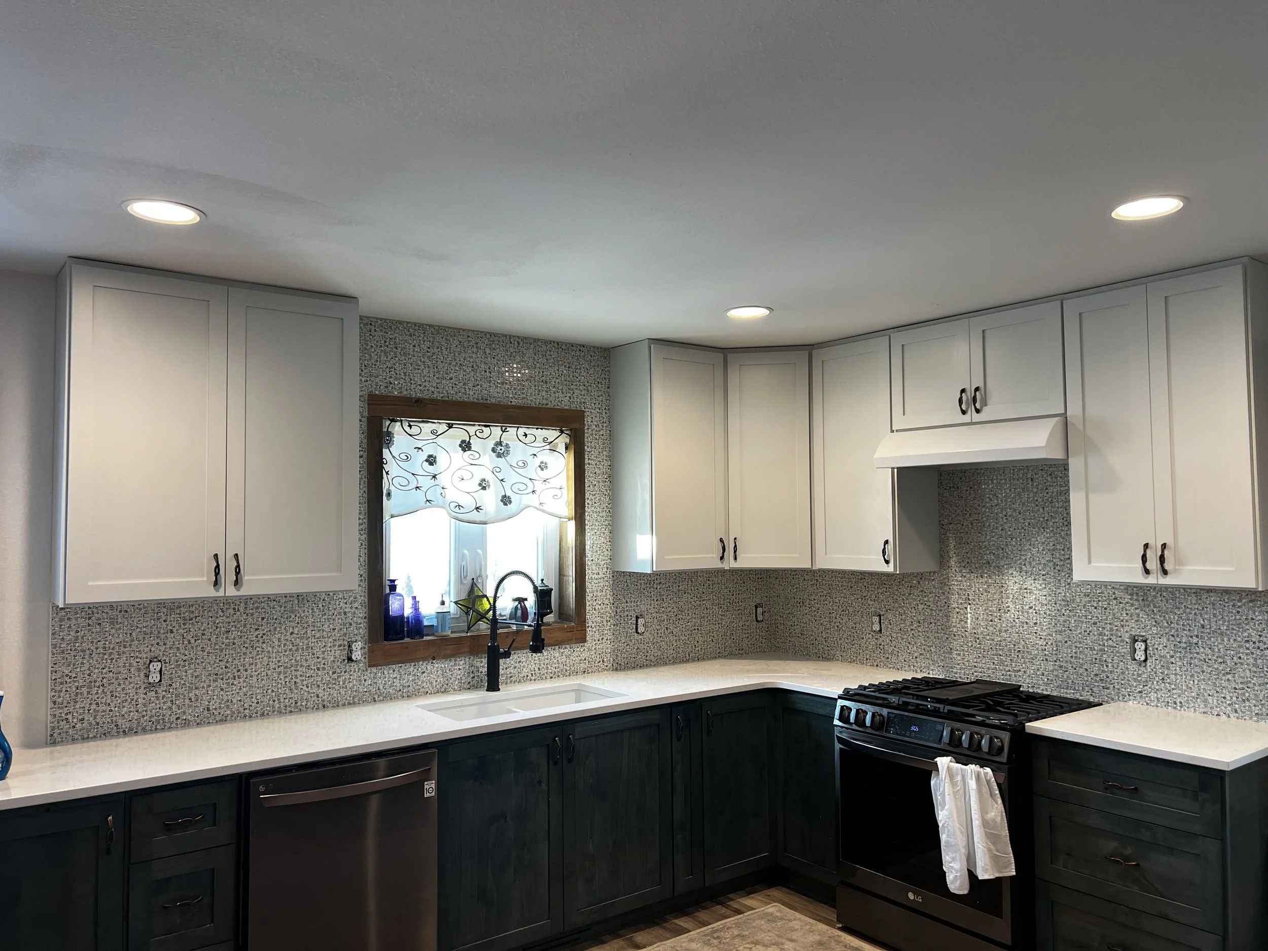Modern kitchen with white upper cabinets, dark lower cabinets, a white countertop, a black stove, and a window with a decorative curtain.