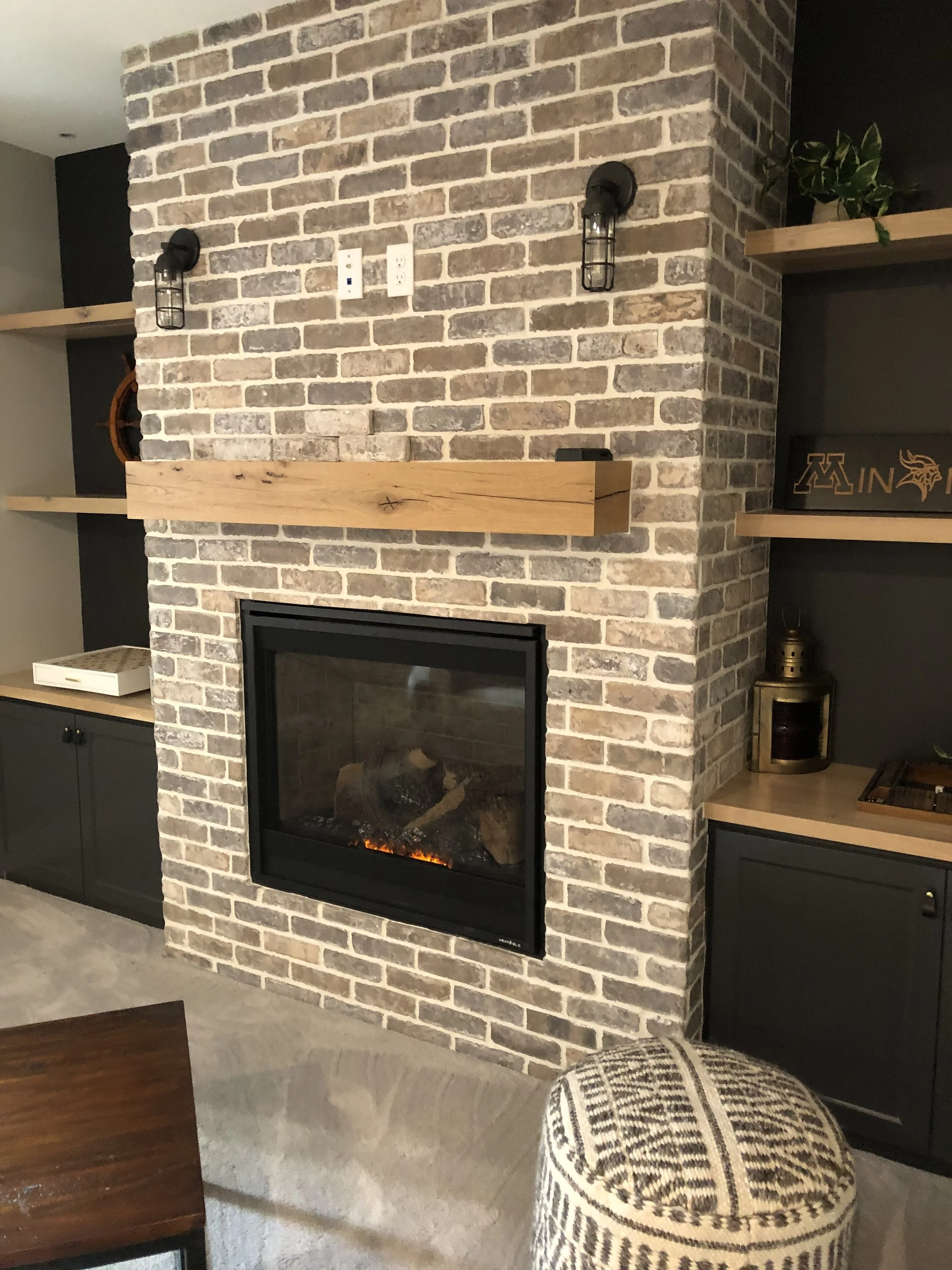 A brick fireplace with a wooden mantel, black wall sconces, and a glass front, surrounded by built-in shelves with decorative items and a plush upholstered stool in front.