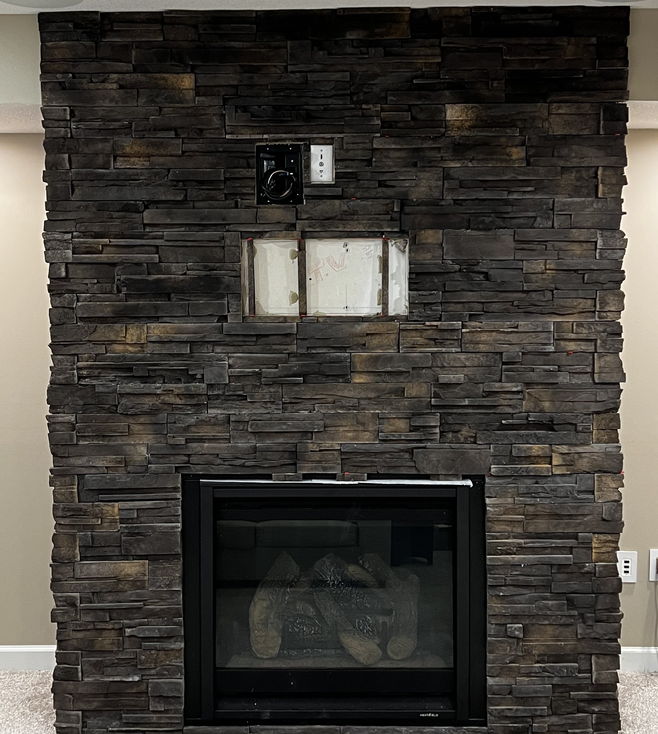 Stone fireplace with a black frame and logs inside, and an unfinished wall above it with a metal box and wiring.