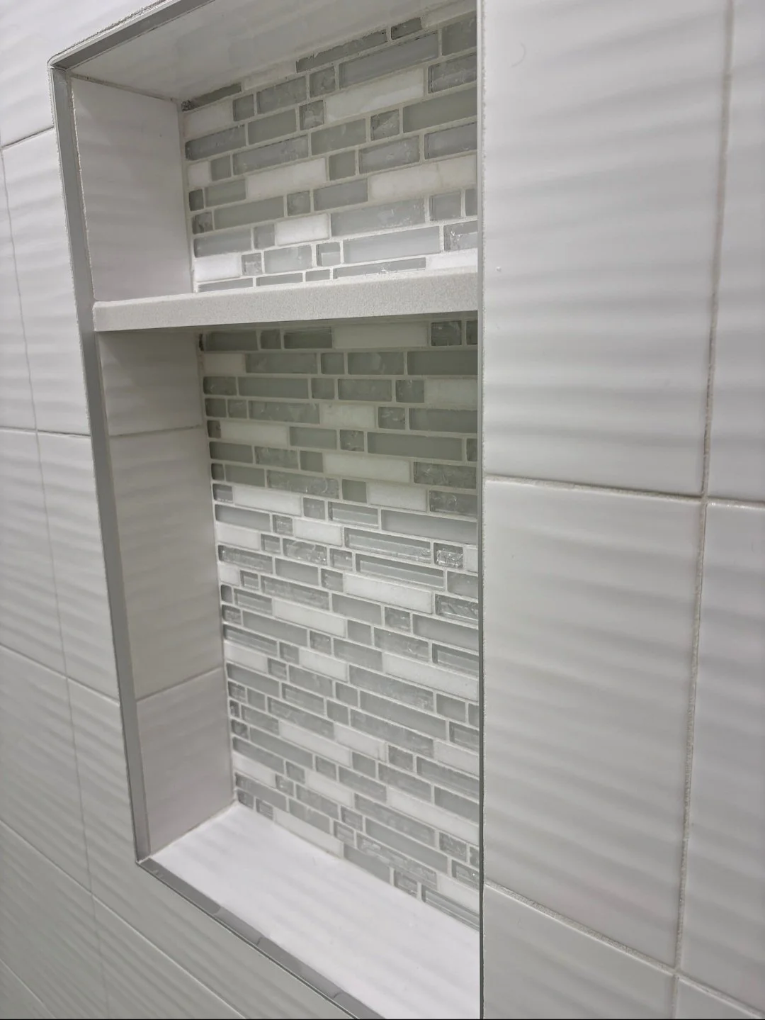 Built-in shower niche with gray and white mosaic tile background, two shelves, and white tiled surround.