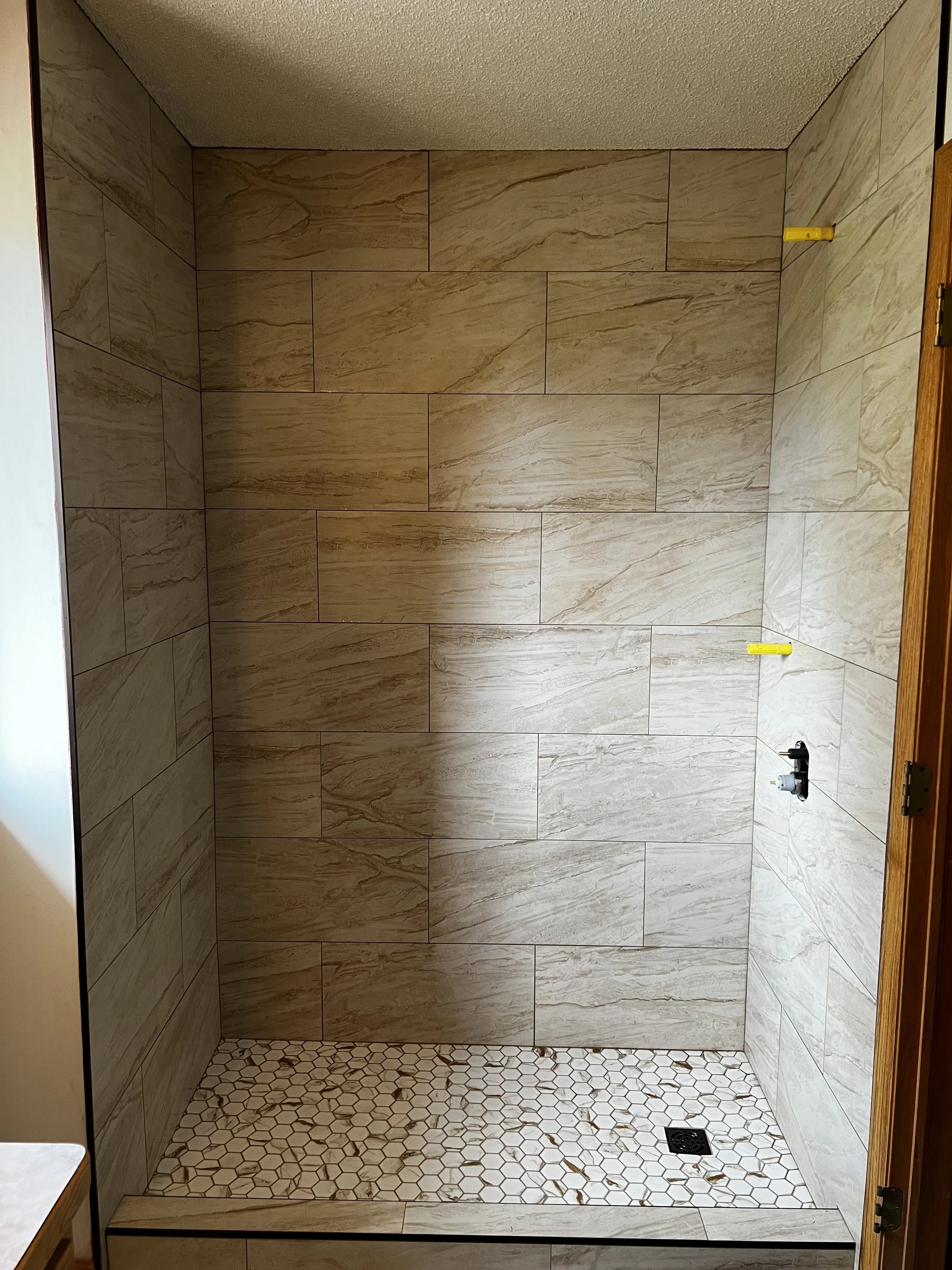 An empty shower area with large beige tiles on the walls and small white hexagonal tiles on the floor, with a drain in the lower right corner.