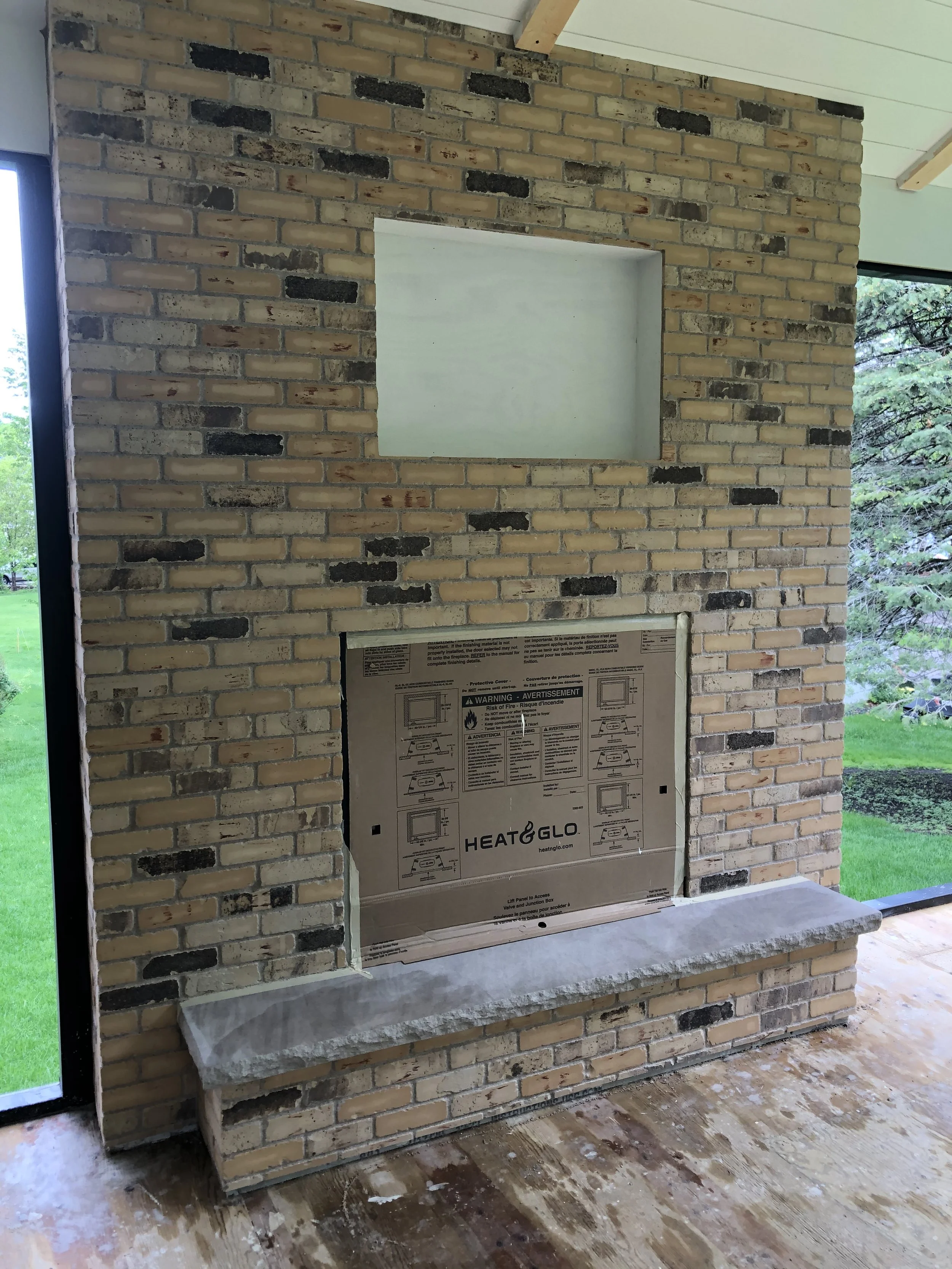 Brick fireplace with an opening covered by protective material, in a room with a wooden floor and large windows showing greenery outside.