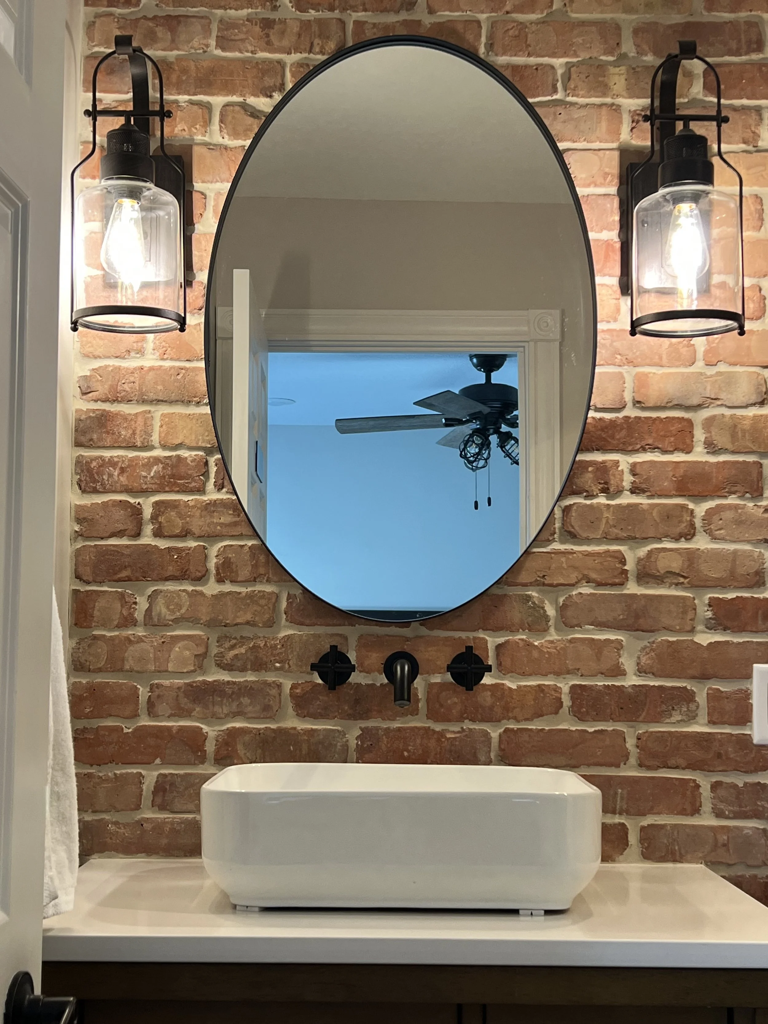 Bathroom vanity with a white vessel sink, black wall-mounted faucet, oval mirror, and industrial-style wall sconces on a brick wall.
