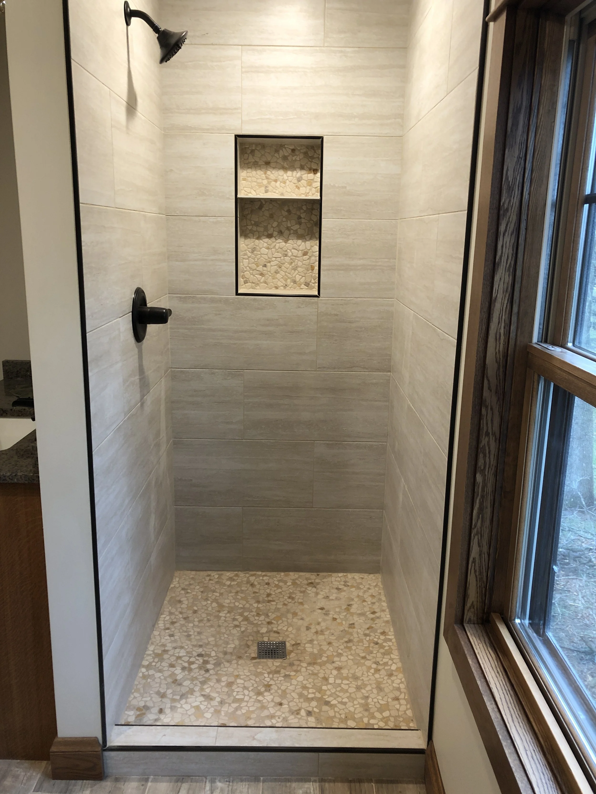 Empty walk-in shower with beige stone tiles on the walls and pebble tiles on the floor, a built-in niche with pebble tiles, a black showerhead, and a window with wooden trim.