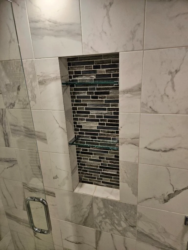 Bathroom shower with marble tiles and a built-in niche with dark brick tiles and glass shelves.