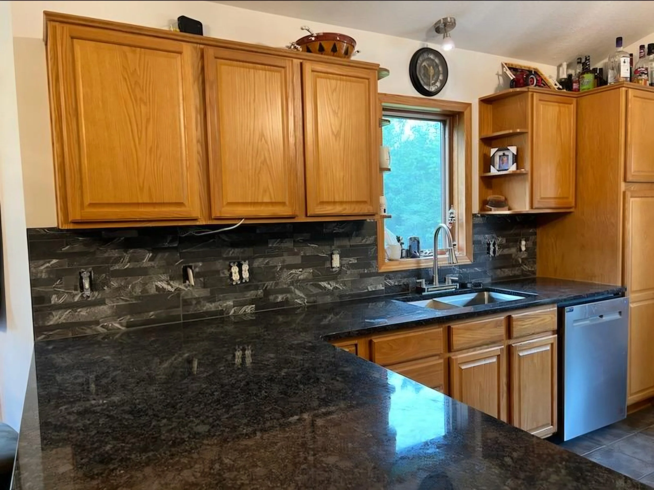 Kitchen with wooden cabinets, black marble countertop and backsplash, stainless steel sink, window above the sink, and a dishwasher.