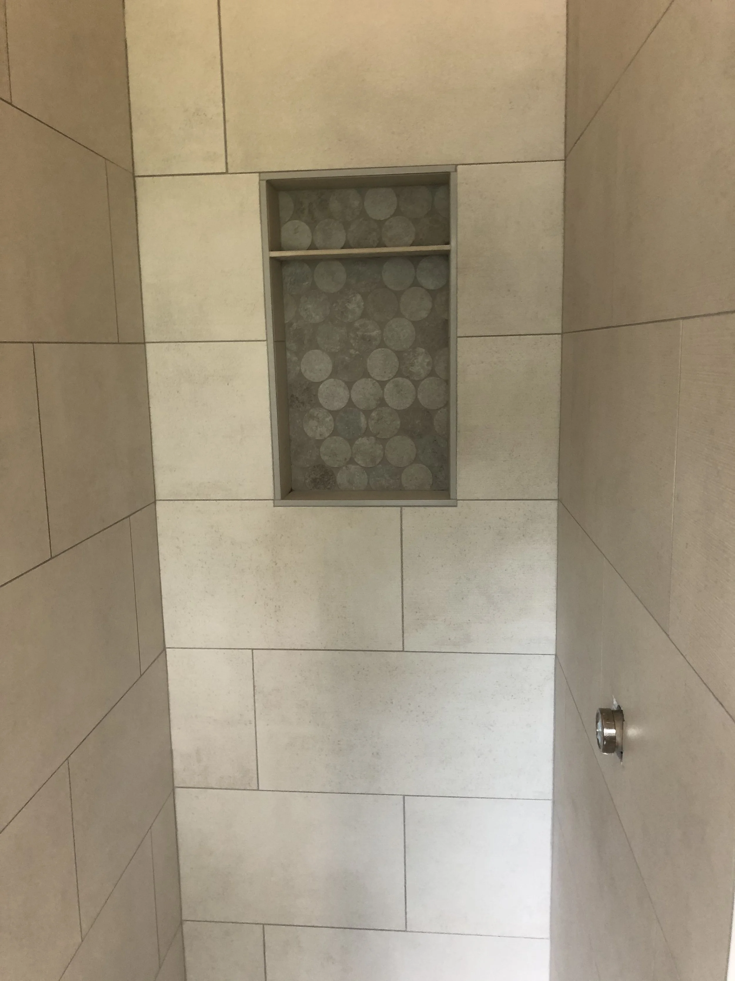 A bathroom shower wall with large beige tiles, a built-in niche with circular pebble tile background, a metallic fixture on the right side.