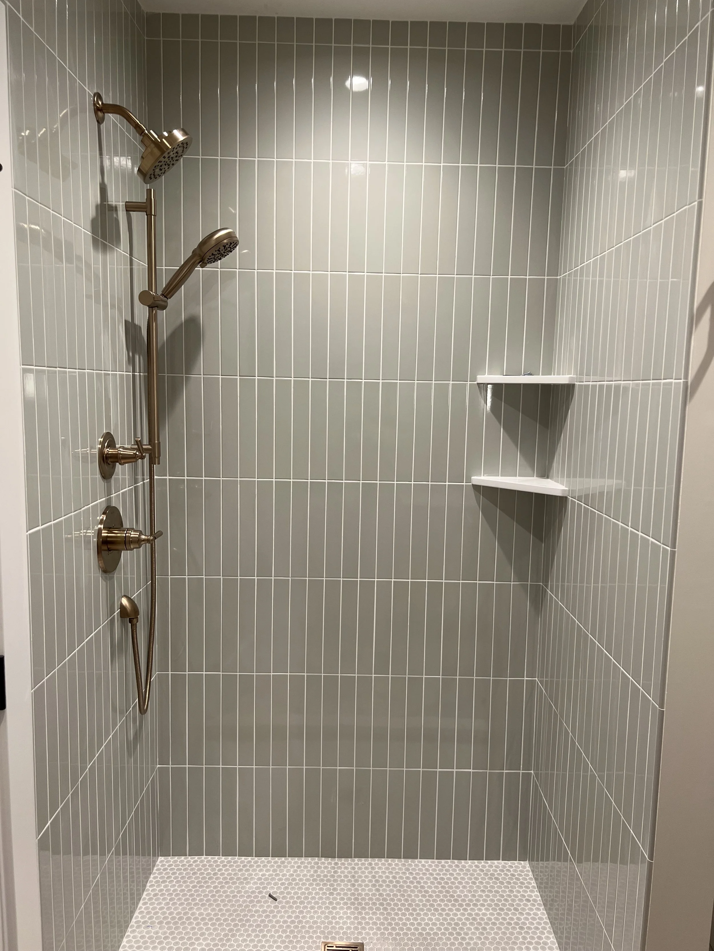 Empty walk-in shower with gray vertical tiles, three white corner shelves, and a brass showerhead and fixtures.