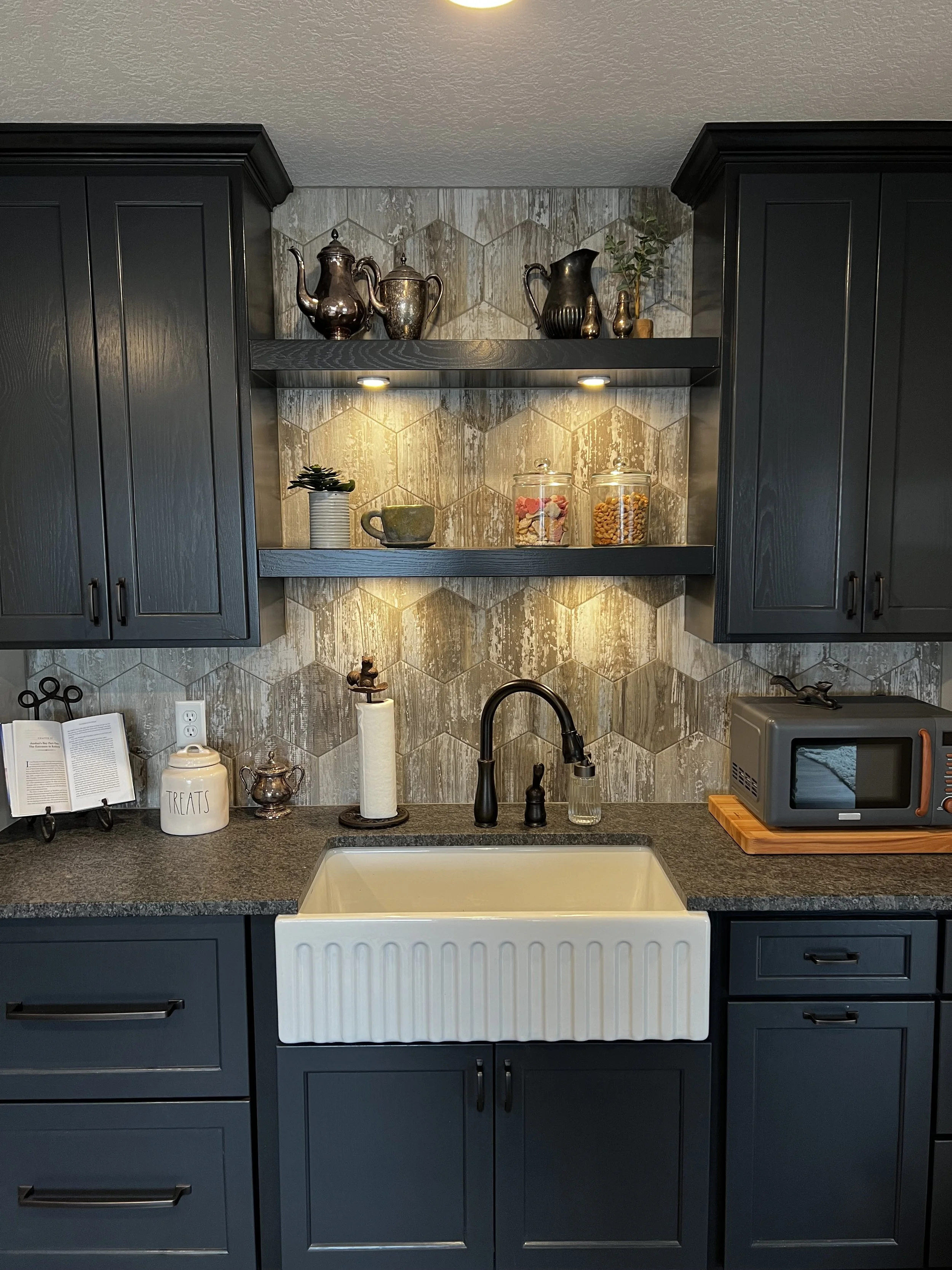 Kitchen with navy blue cabinets, a white farmhouse sink, dark countertop, open shelves with teapots, jars, plants, and decor, a microwave, and hexagonal tile backsplash.