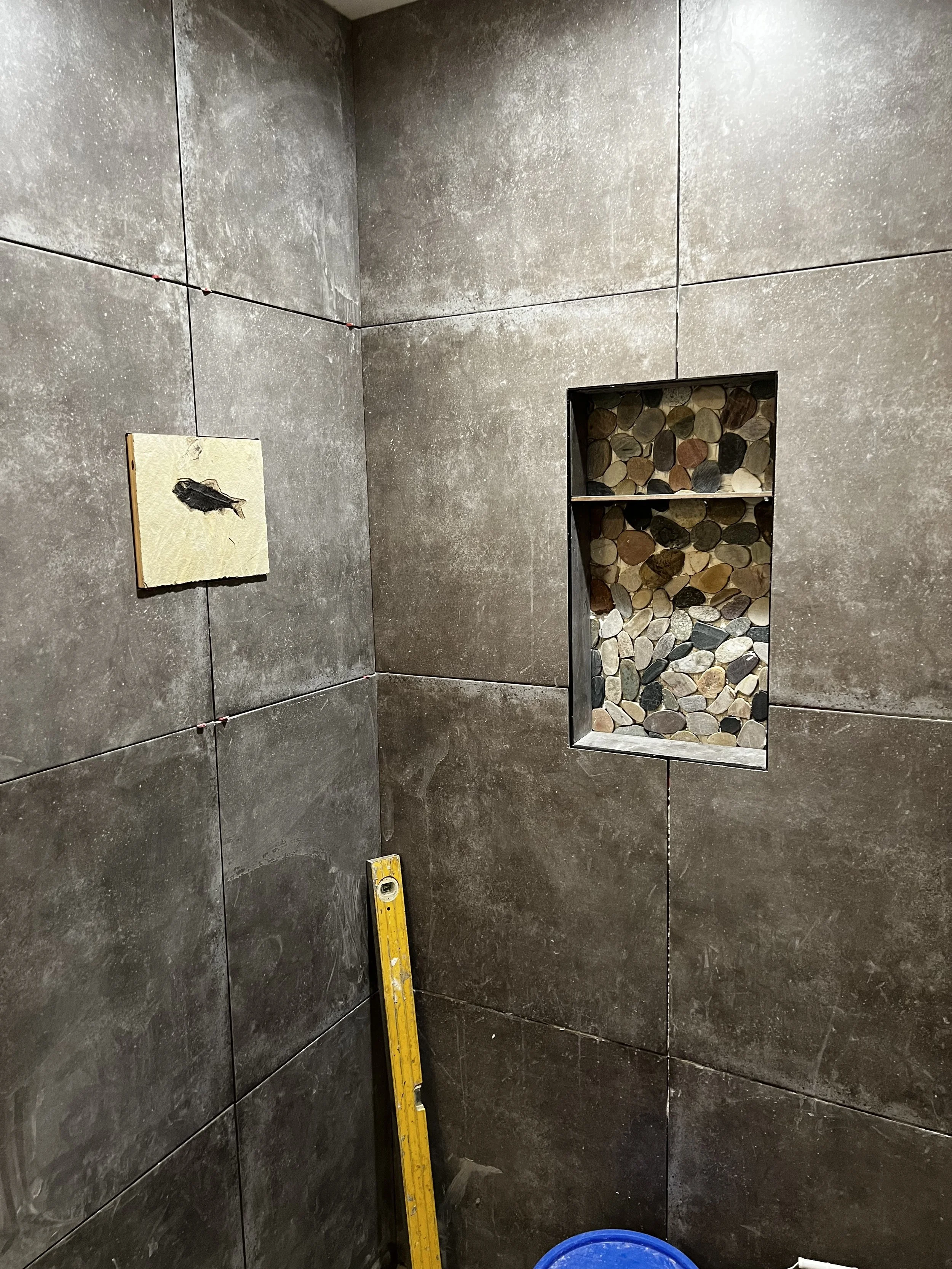 The image shows a tiled bathroom corner with large gray tiles, an embedded niche filled with multicolored smooth stones, a small piece of drywall, a yellow level leaning against the wall, and a blue bucket on the floor.
