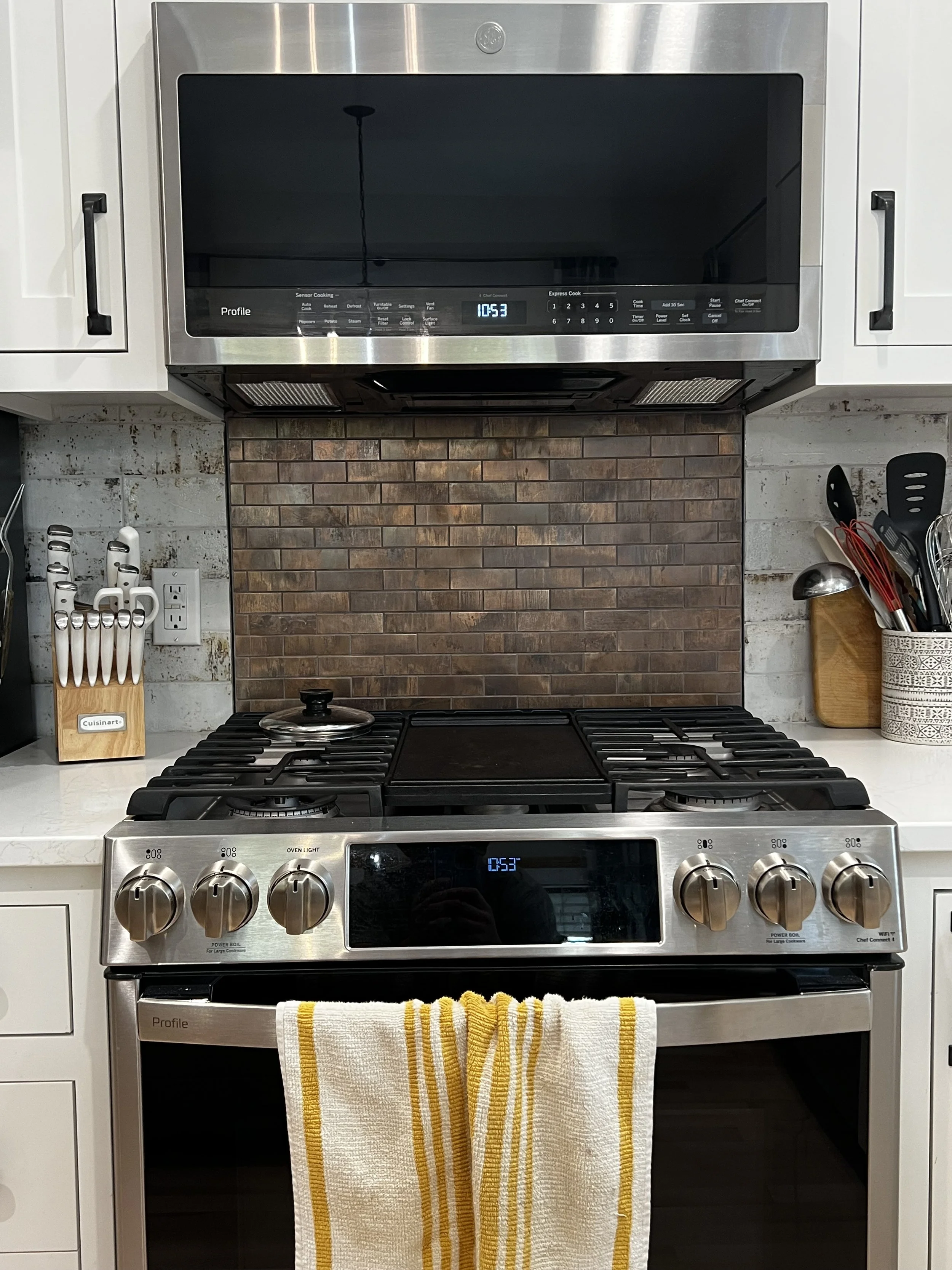 A modern kitchen stove and microwave with a brick backsplash. The stove is stainless steel with five knobs and a towel hanging on it. The microwave is mounted above the stove and shows the time 10:53.