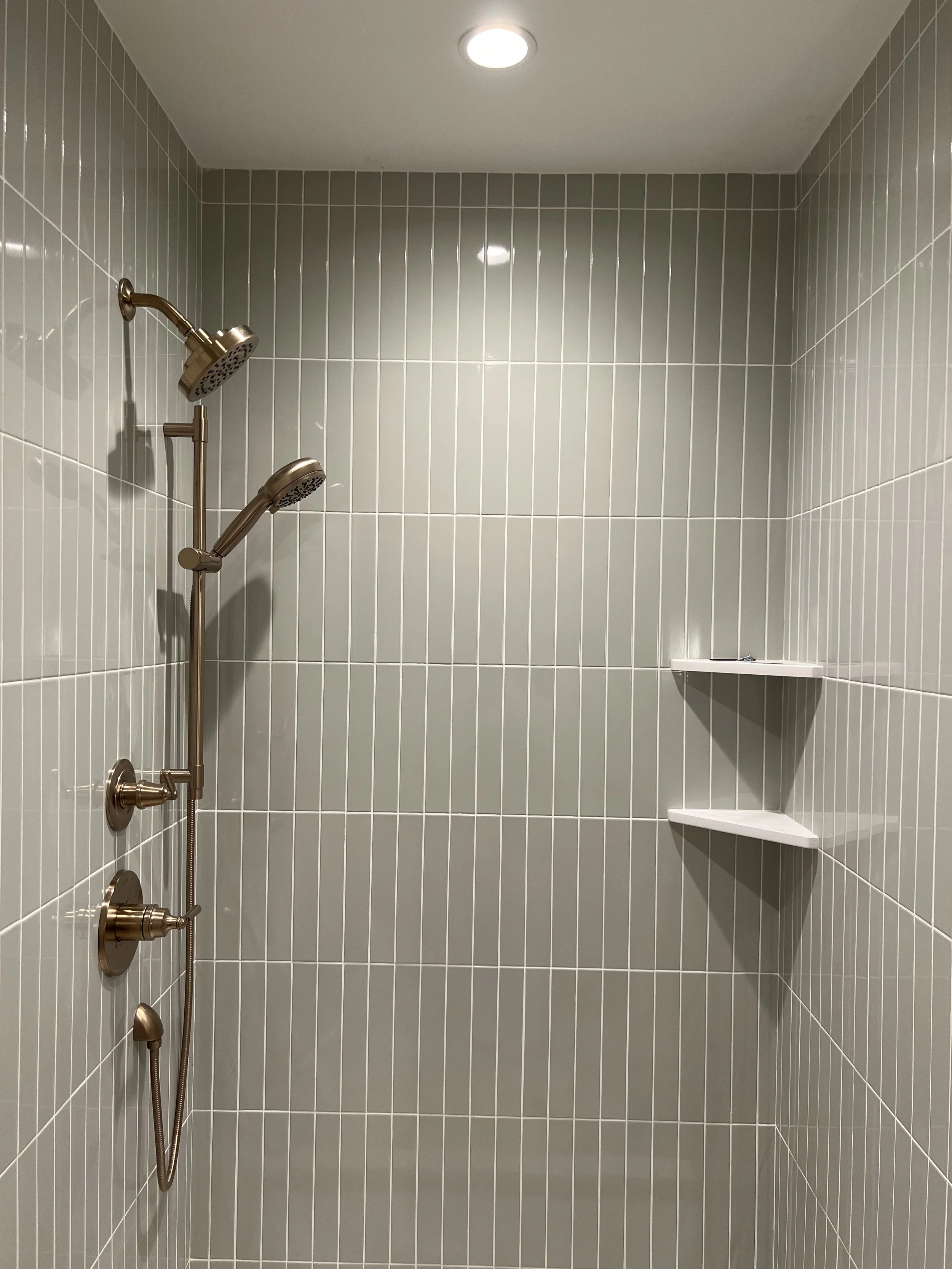 Empty shower with gray tiled walls, two white corner shelves, and a ceiling light. A dual showerhead system with brushed nickel finish is attached to the left wall.