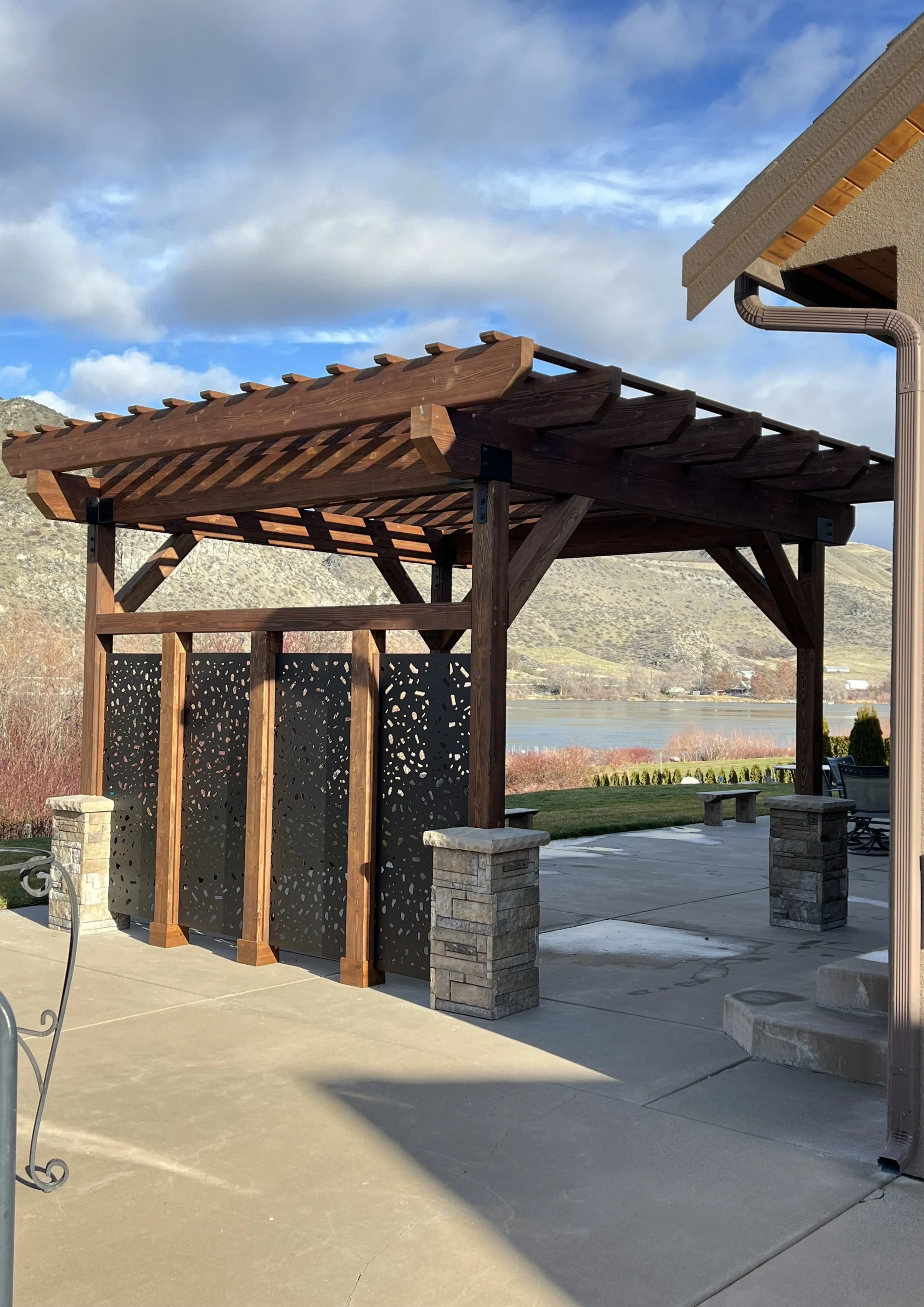 A wooden pergola with a decorative privacy screen on a concrete patio, overlooking a scenic view of a lake and mountains under a partly cloudy sky.