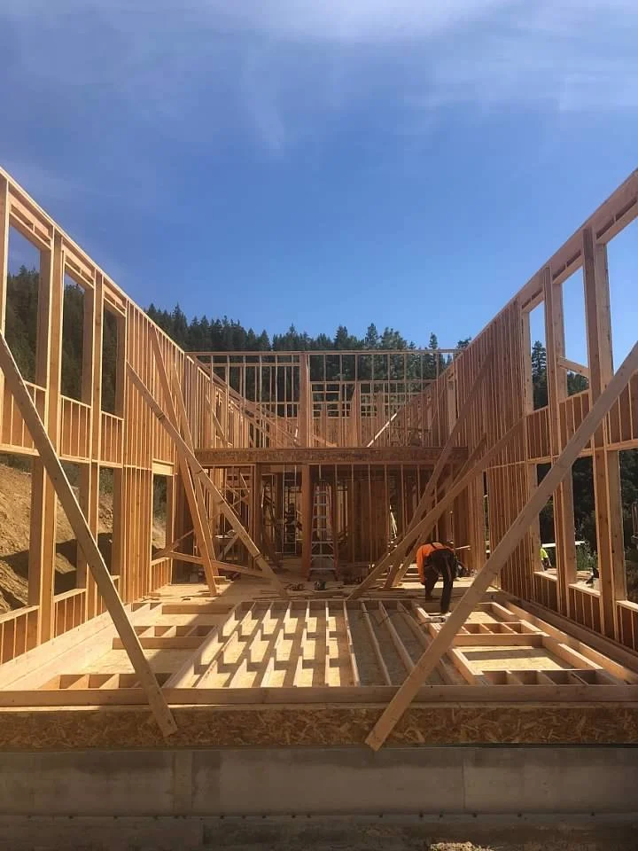 Construction workers build the wooden framework of a house during daylight, with a clear blue sky and trees in the background.