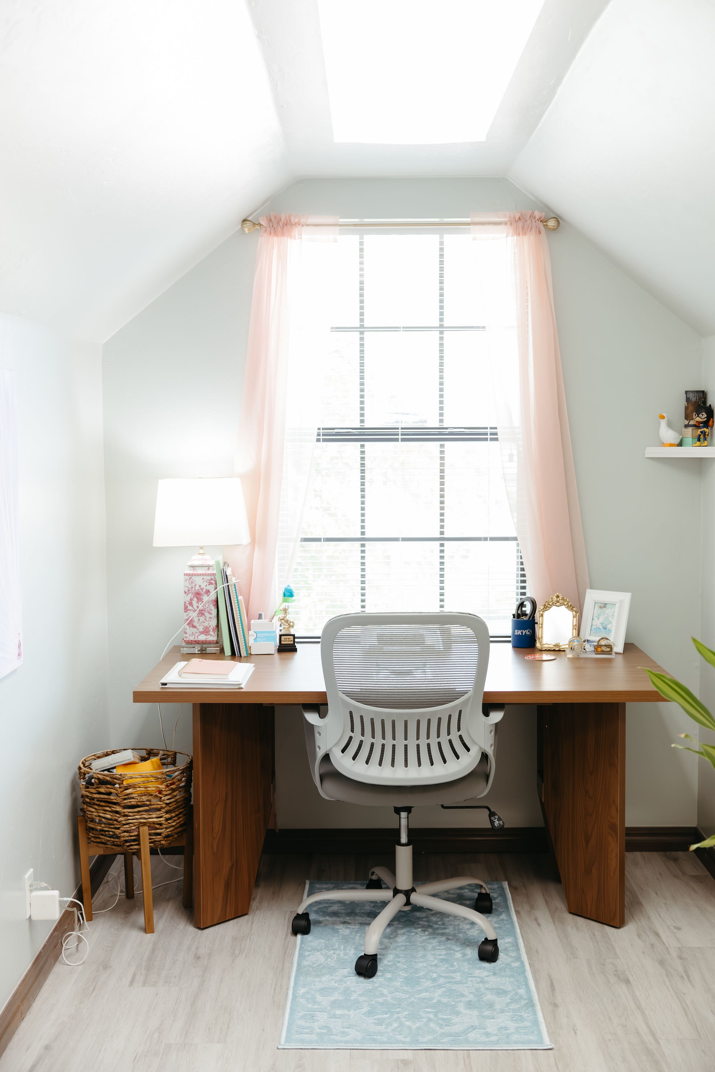Therapy office with a wooden desk, a white ergonomic chair, and decor including a lamp, framed photo, and stationery, in a room with a large window, pink curtains, and skylight.