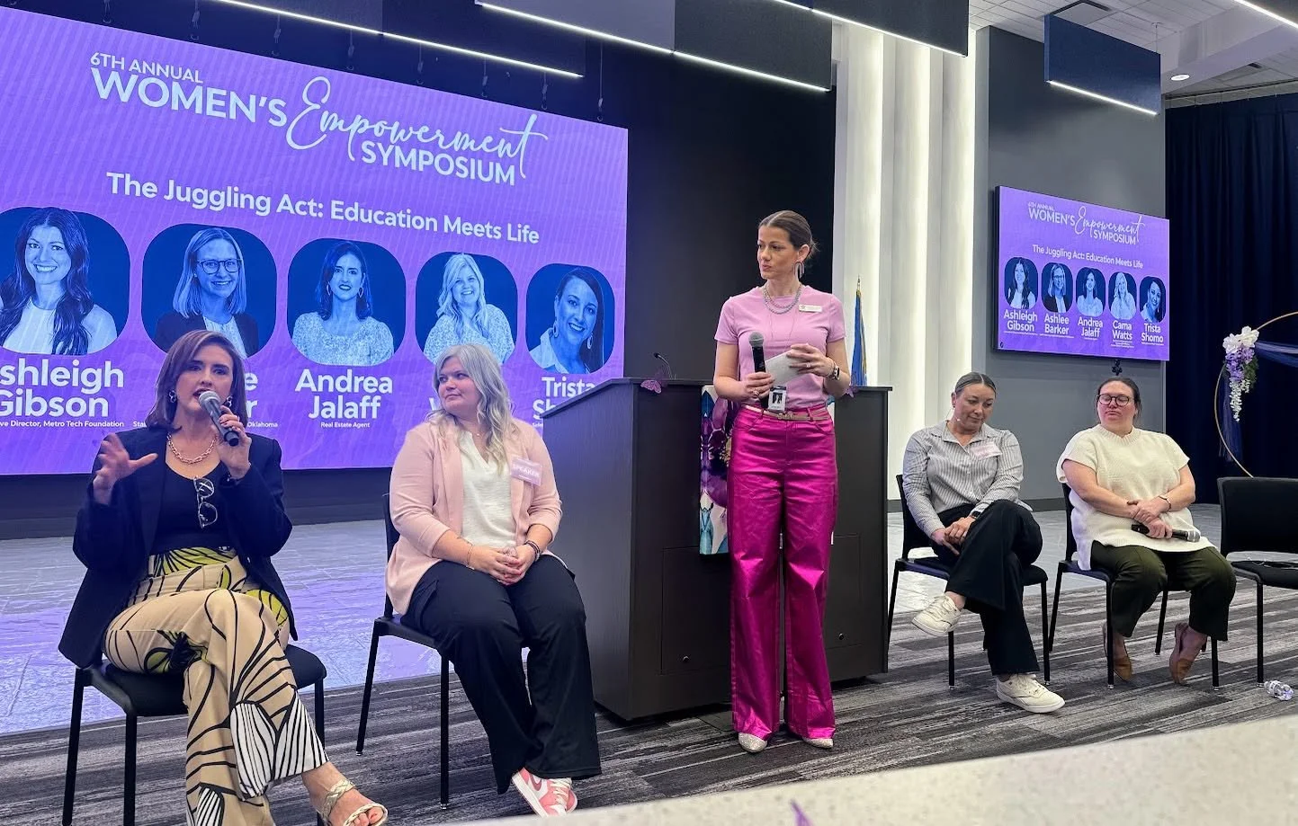 Woman moderating a panel during the 6th Annual Women's Empowerment Symposium with a large screen behind them displaying the event title, speakers' names, and photos.
