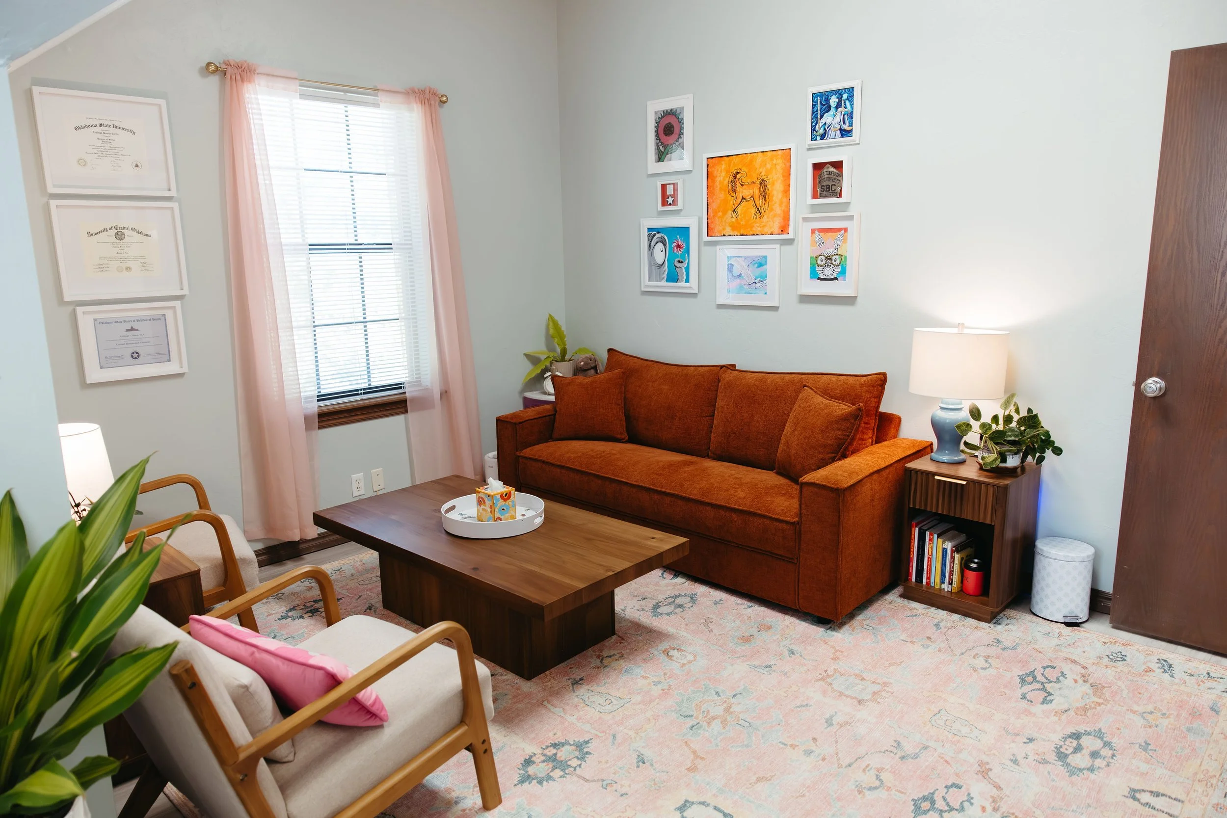 A cozy living room with a brick-colored sofa, a wooden coffee table with tissues, a side table with a lamp, framed artwork on the wall, a window with pink curtains, and a cream-colored armchair with pink pillow. There are houseplants and a patterned 