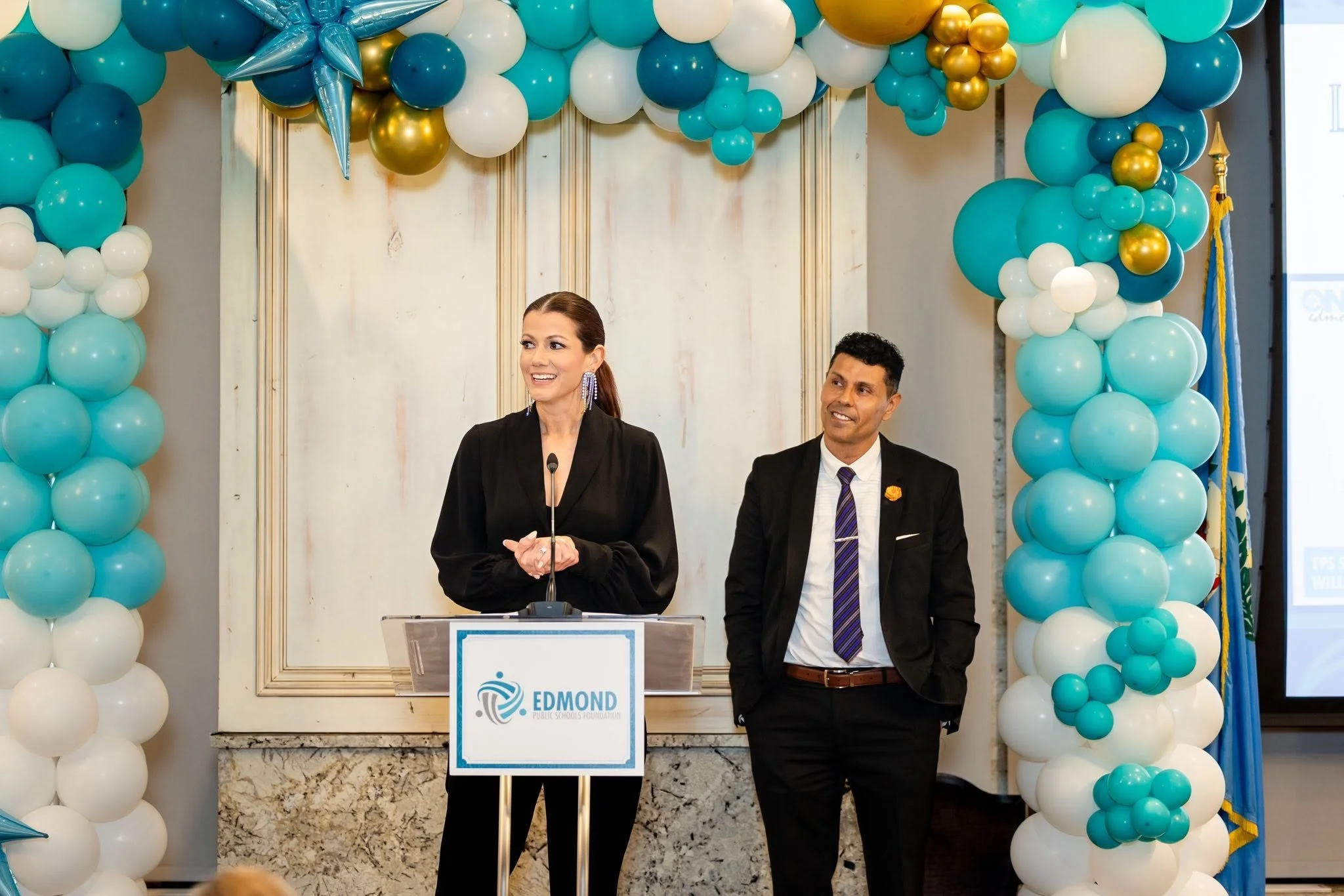 Two individuals standing at a podium amid balloon decorations in shades of blue, white, and gold, in a formal indoor setting.