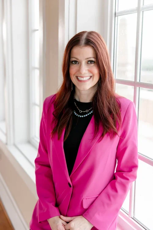 A woman with long red hair wearing a bright pink blazer and a black shirt, standing by a window with white framing, smiling at the camera.