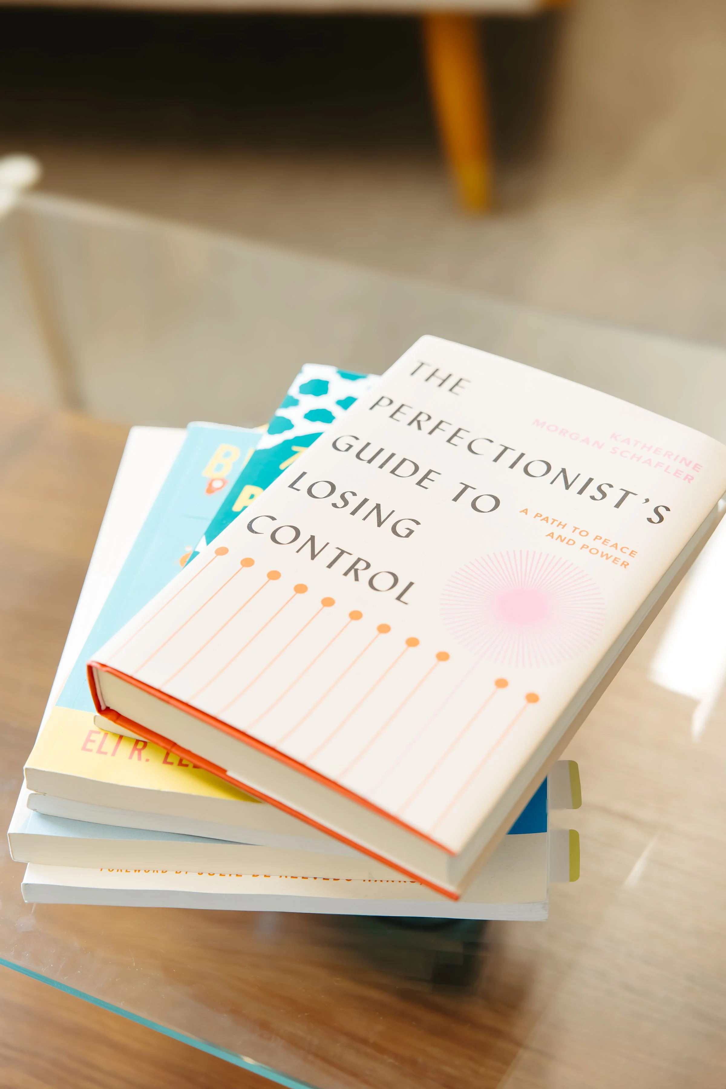 Multiple psychology books stacked on a wooden table, with the top book titled 'The perfectionist's guide to losing control' by Katherine Morgan Schafler.