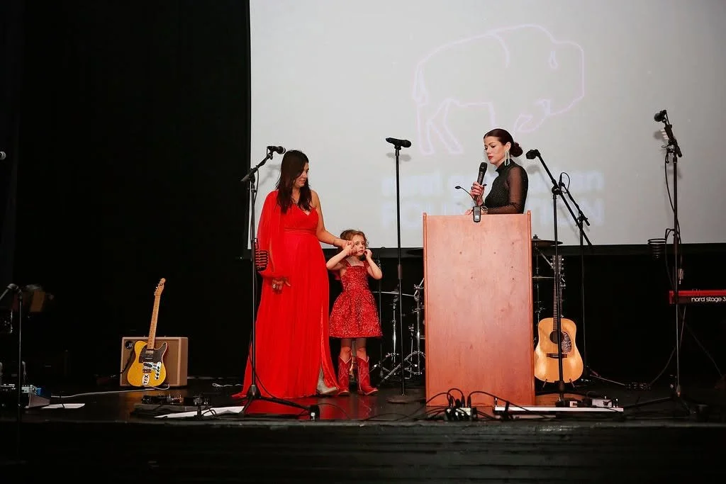 A woman in a red dress, a young girl in a red dress, and another woman in black are on stage with microphones, guitars, and a drum set, during a performance or presentation.