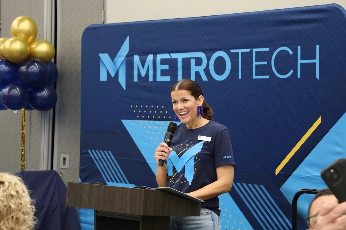 A woman with brown hair, blue earrings, and a navy blue Metro Tech t-shirt, smiling and speaking into a microphone at a Metro Tech event. She is standing at a podium with a blue and yellow themed backdrop and balloons in the background.