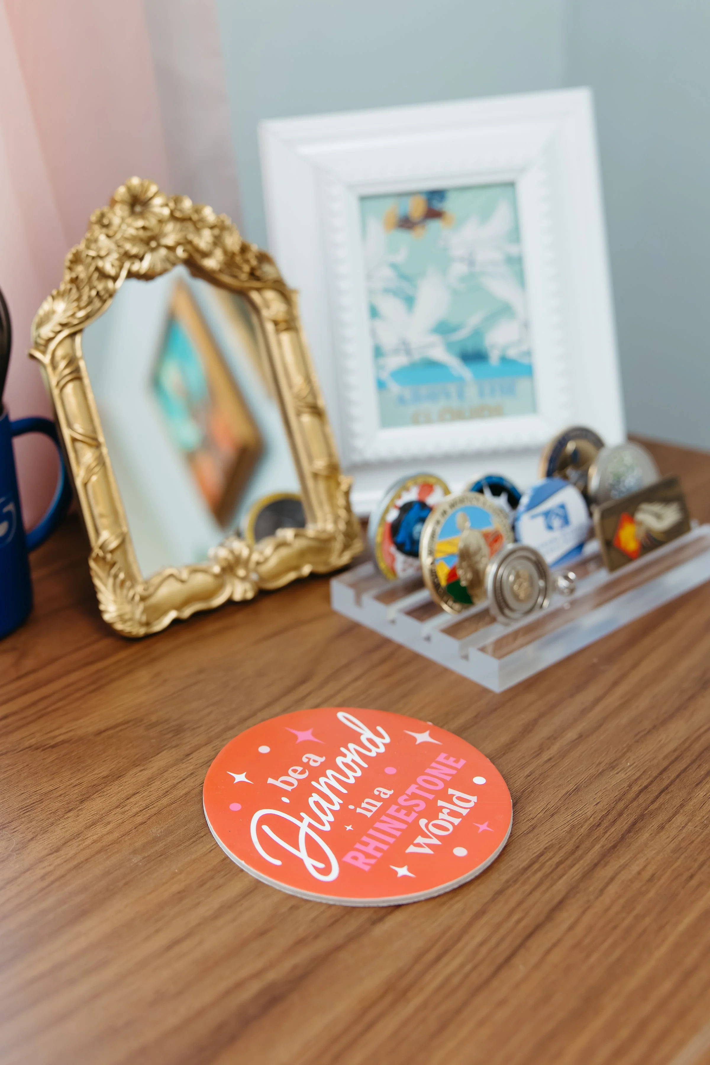 Decorative desk setup with ornate gold mirror, framed artwork, and various collectible pins or badges on a white stand, with an orange circular sticker on a wooden surface in the foreground.