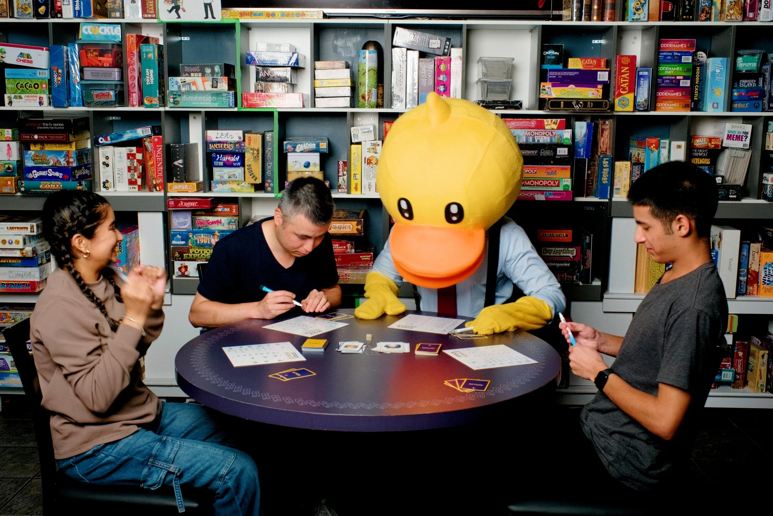 Four people playing a board game at a round table, one person dressed in a duck costume, in a room with shelves of board games.