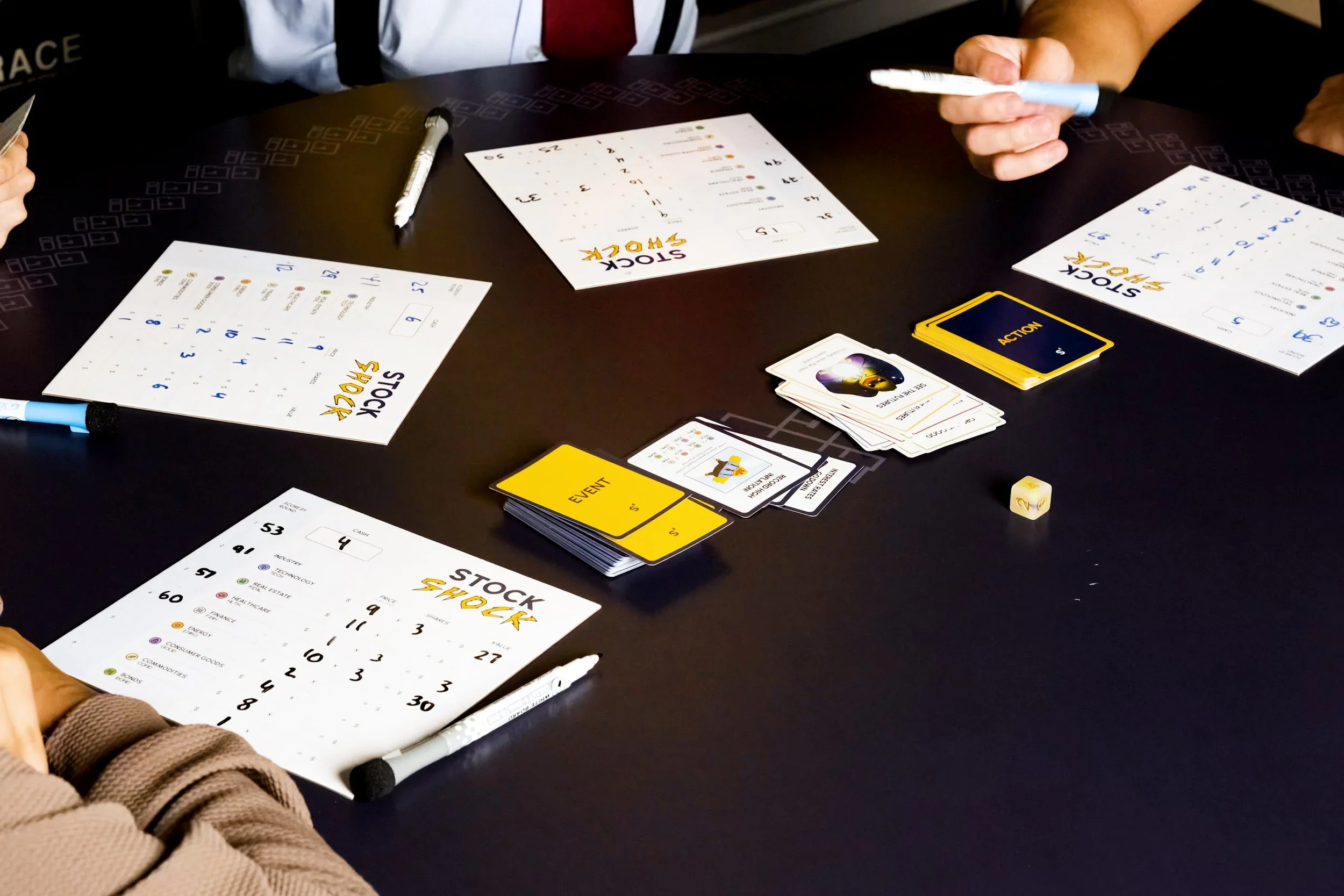 People playing a board game called Stock Exchange with paper sheets, pens, action and event cards, and dice on a black table.