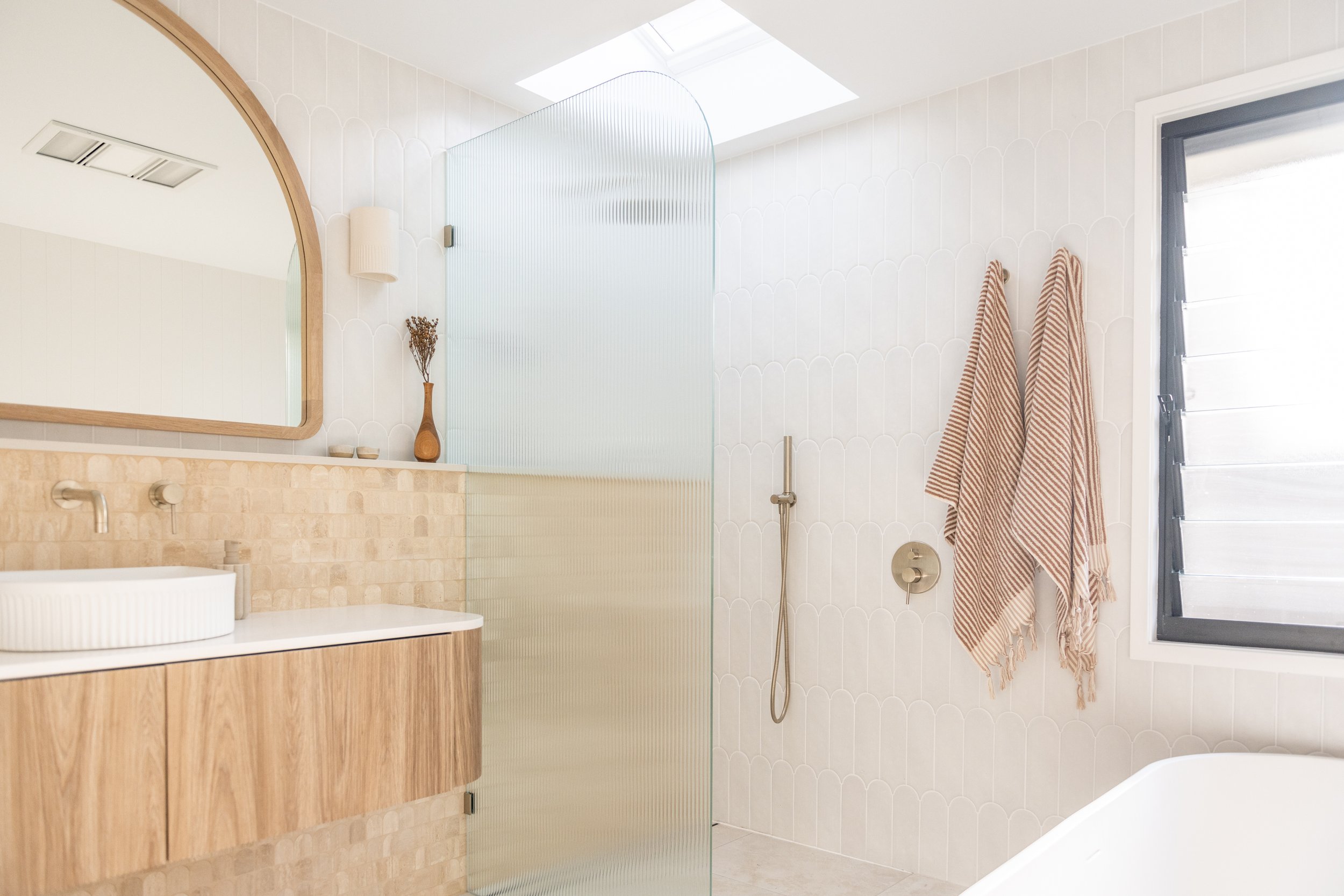 Modern bathroom with a wooden vanity, white vessel sink, large oval mirror, beige tiles, a frosted glass divider, wall-mounted showerhead, two striped towels hanging on the wall, a window with adjustable slats, and a skylight.
