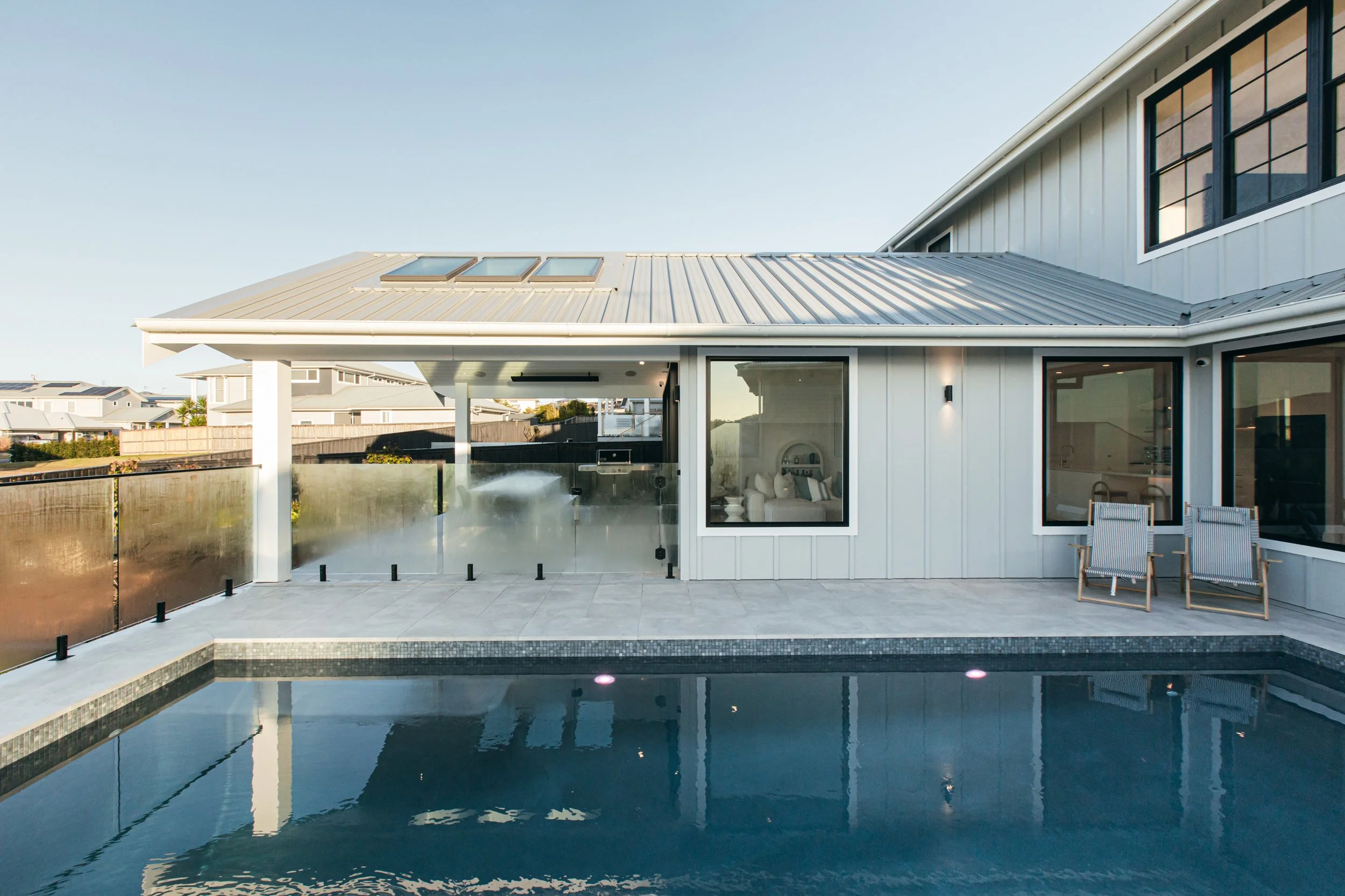 Modern house with a stucco exterior, large windows, and metal roofing, featuring a backyard with a swimming pool, two lounge chairs, and a glass privacy fence surrounding the pool area.