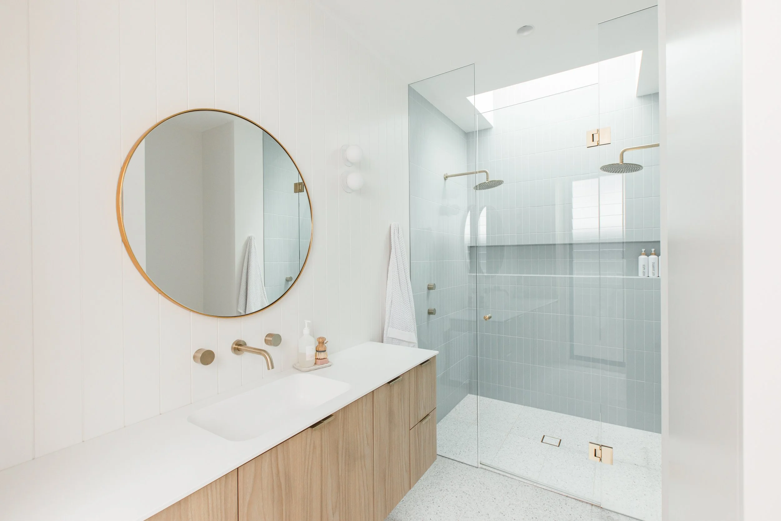 Modern bathroom with white walls, a round mirror, a light-colored wooden vanity, and a glass-enclosed shower area with two showerheads and built-in shelves.