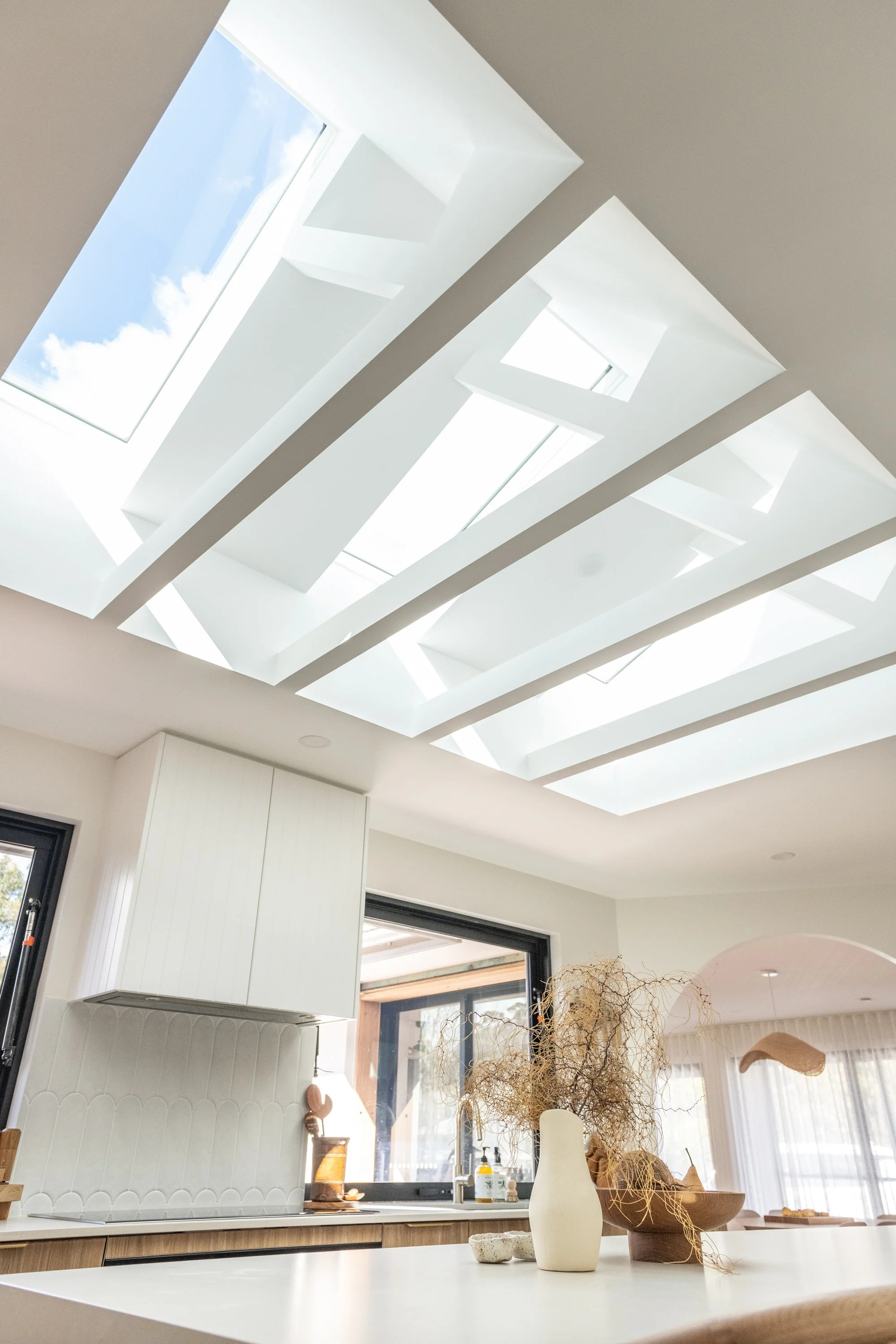 Modern kitchen with skylights in the ceiling providing natural light, featuring a white countertop, decorative vases, and lush plants.