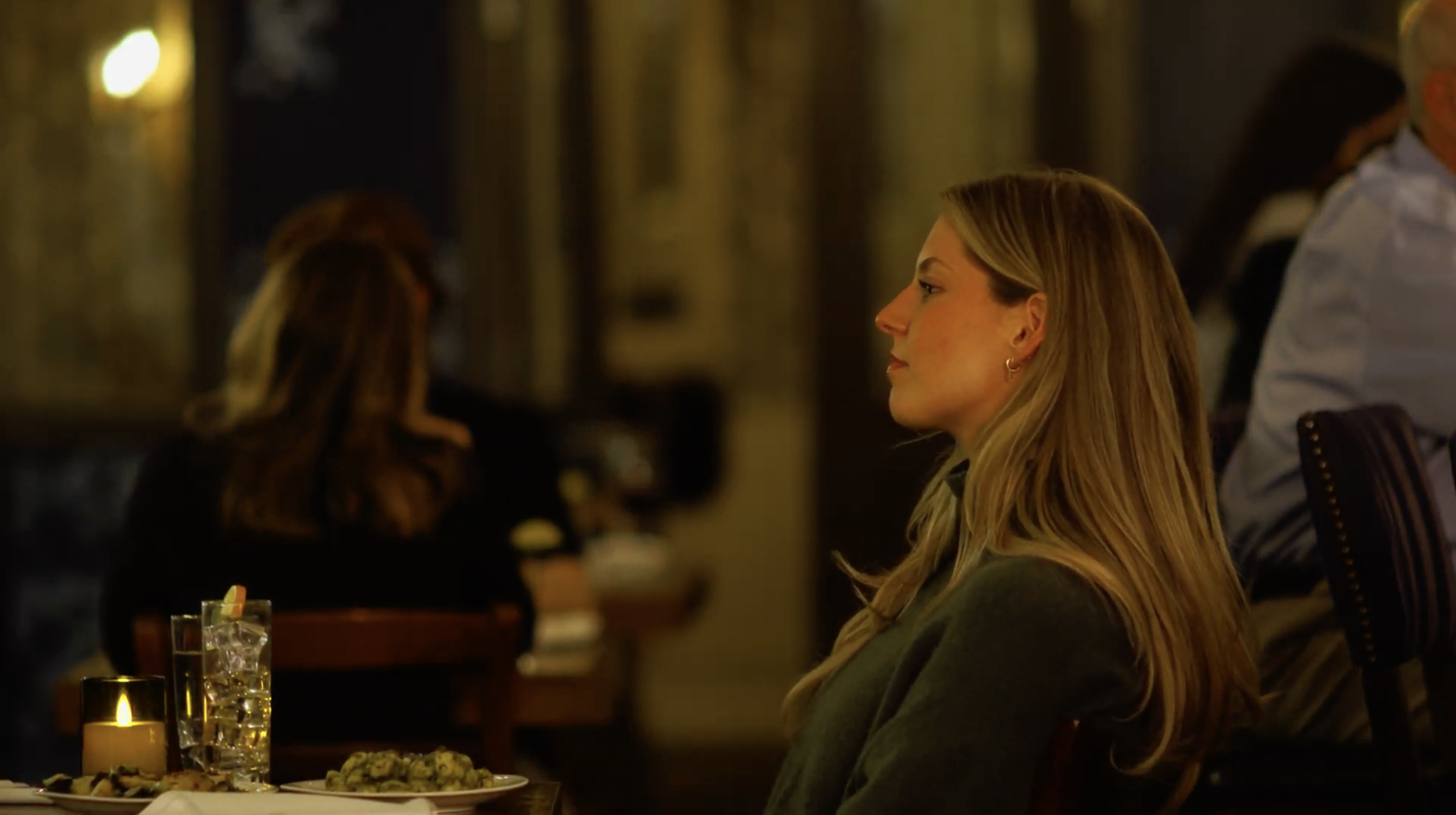 A woman with long blond hair sitting at a table in a restaurant or bar, with food and a lit candle on the table. She is facing right, with a side profile visible. The background is dimly lit with blurred patrons.