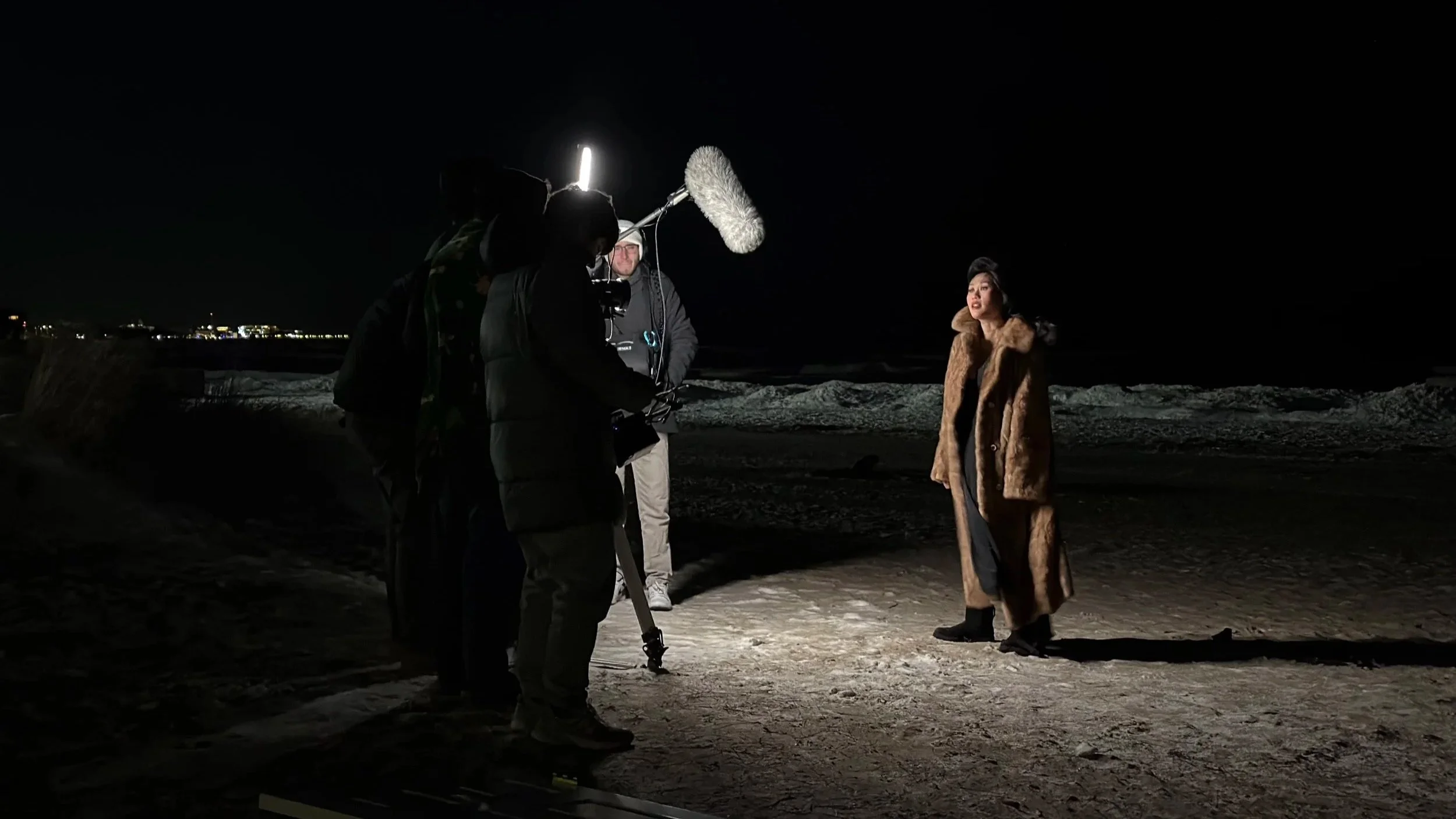 A woman in a long fur coat being filmed during a nighttime outdoor interview or scene on snow or ice, with film crew and equipment in the background.