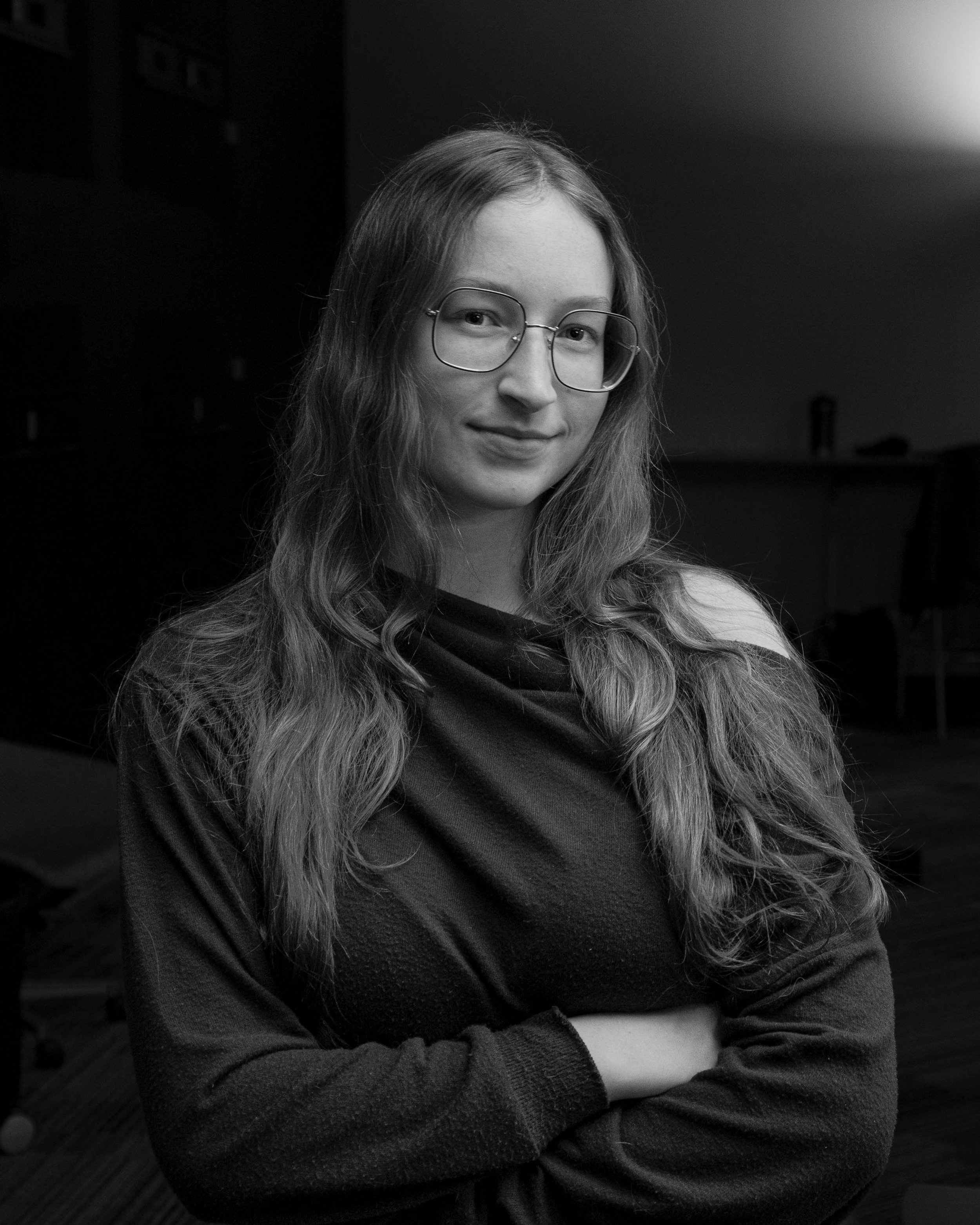 Black and white photo of a young woman with long wavy hair and large glasses, smiling slightly, standing with arms crossed in a dimly lit room.