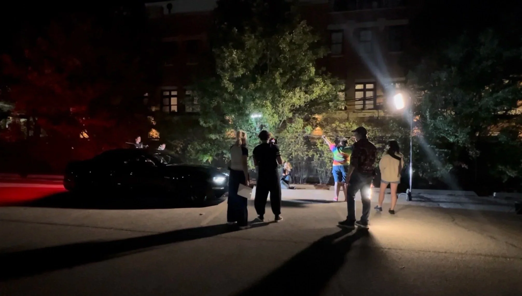 A group of people standing on a street at night, with a black sports car, illuminated by a bright streetlight, and a person holding a colorful umbrella near a tree.