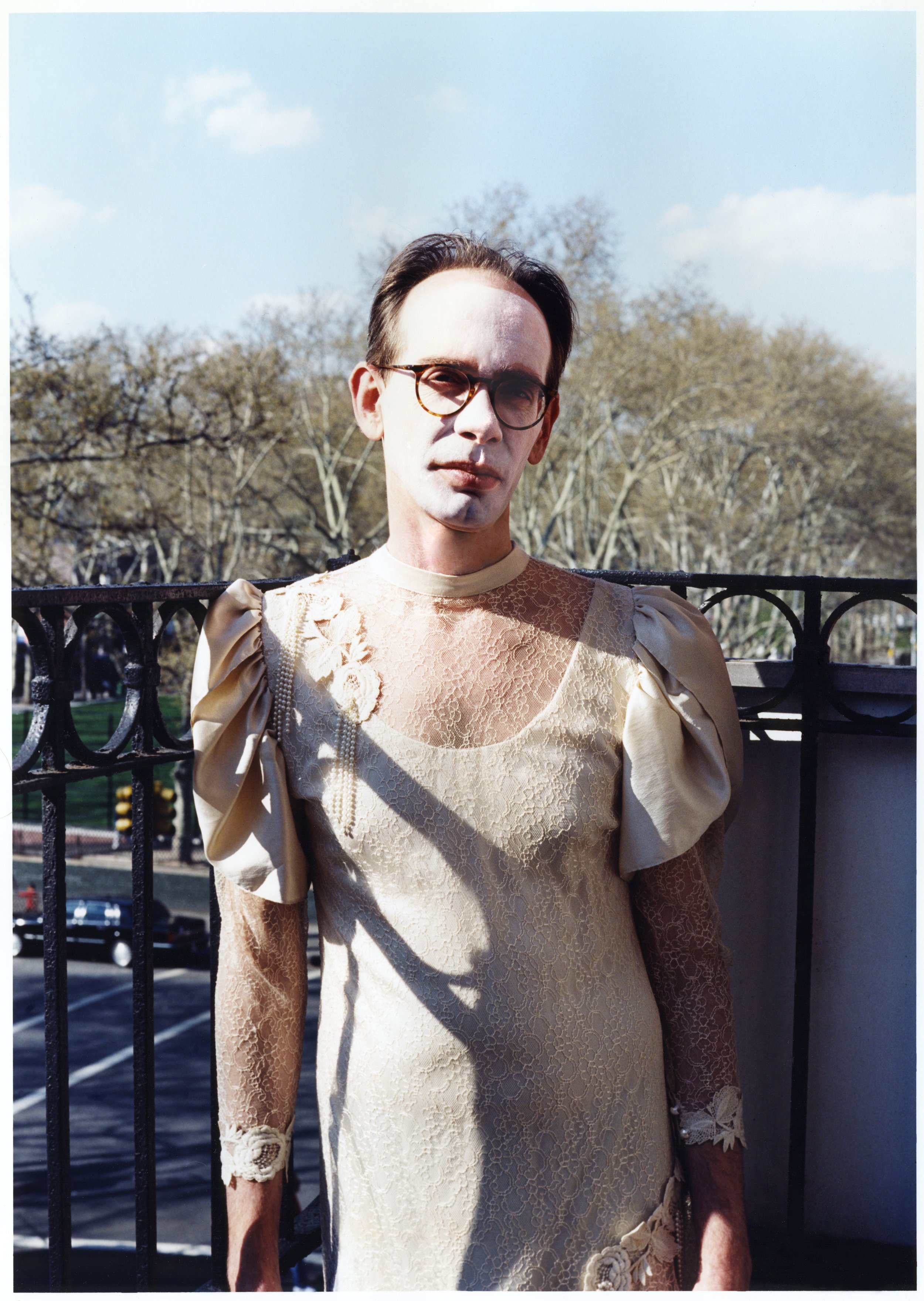 Arto Lindsay dressed in a vintage lace dress with puffed sleeves, standing on a fire escape in Chinatown, NYC, with a park and trees in the background, wearing glasses and makeup.