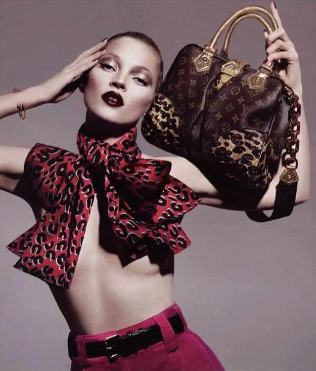 Kate Moss with bold makeup holding a Louis Vuitton handbag and wearing a red and black animal print scarf, pink belt, and pink pants against a plain background.