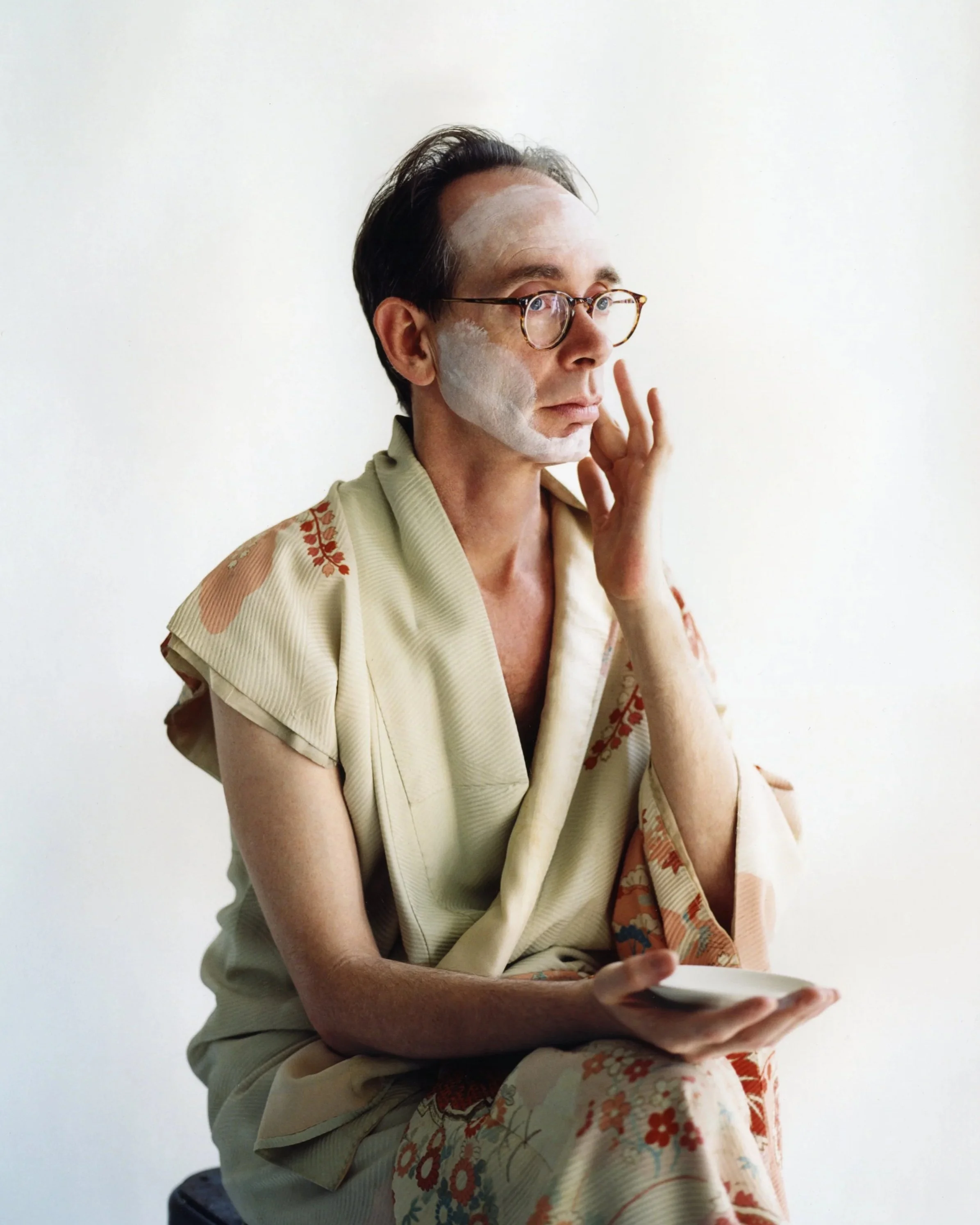 Musicuian Arto Lindsay with glasses sitting on a stool, applying white face makeup and holding a makeup palette, dressed in a kimono with floral patterns, against a plain white background.