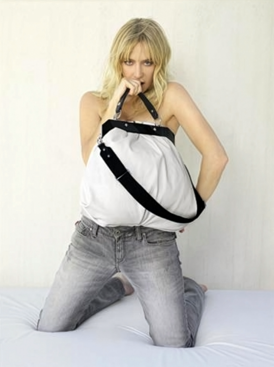 A woman with blonde hair kneeling on a bed, holding a large white bag with black straps and biting the handle.
