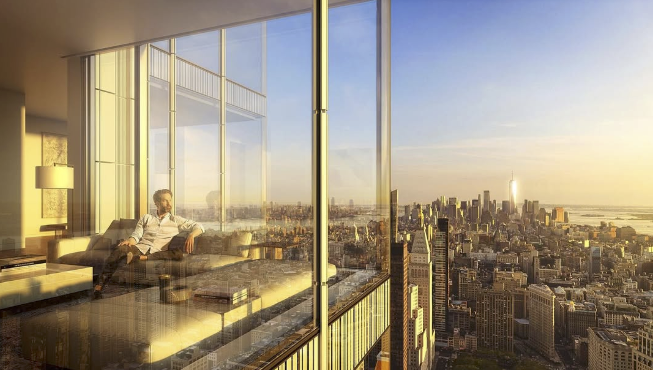 A man sitting on a beige sofa inside a high-rise apartment with floor-to-ceiling windows, overlooking a city skyline during sunset.