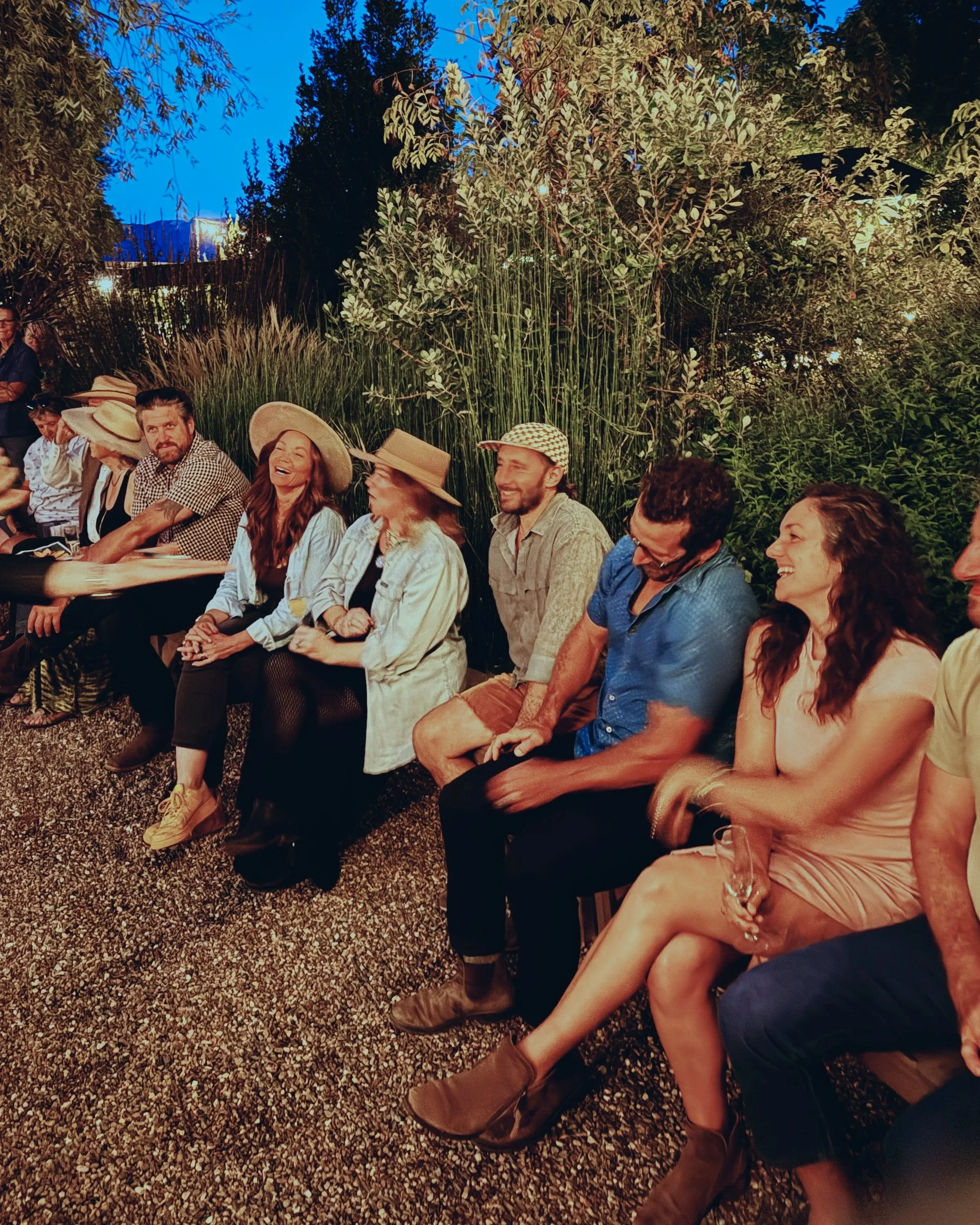 Group of people sitting outdoors at night, engaged in conversation and laughing, with greenery and trees in the background.