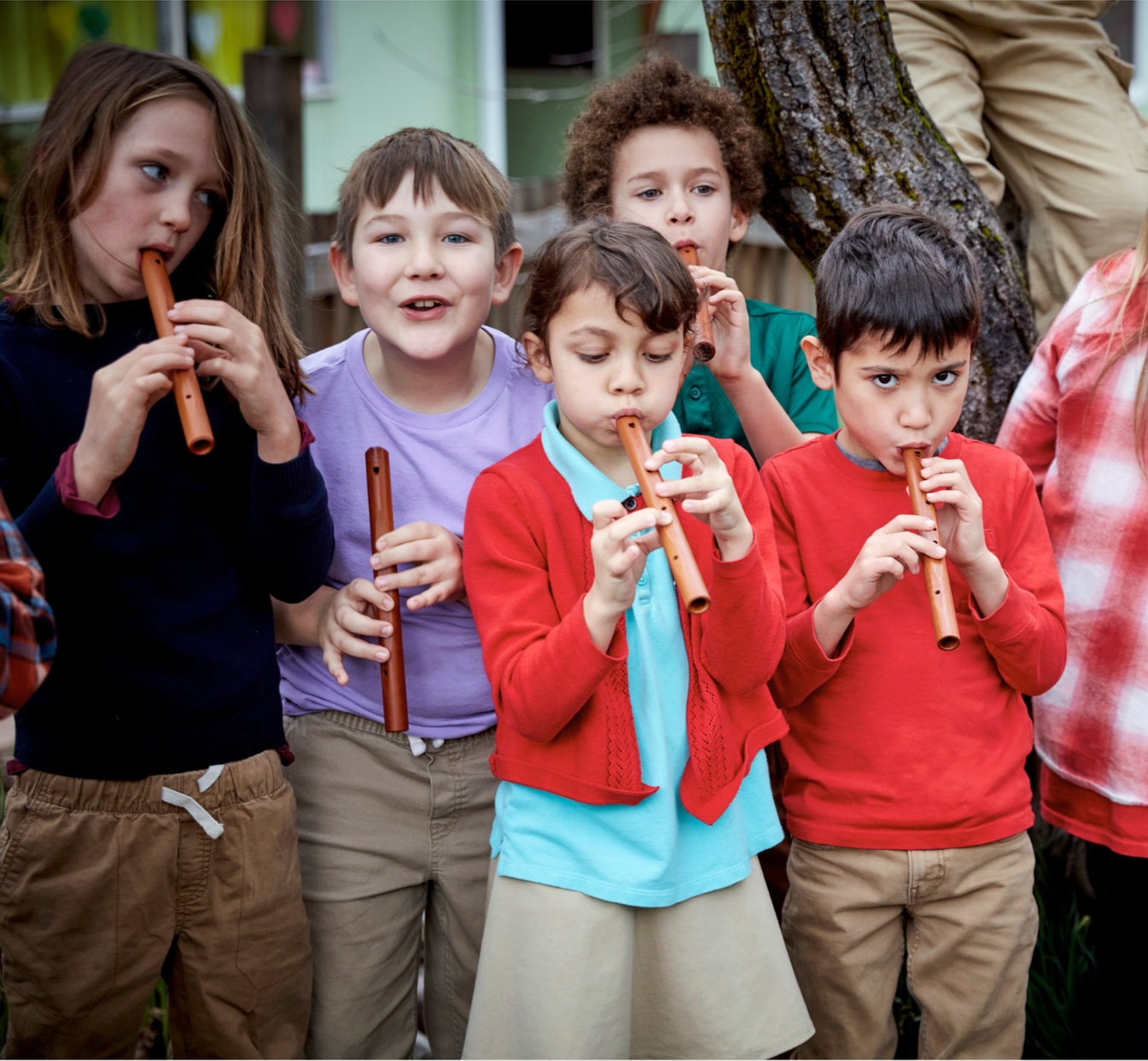 Group of children playing recorders outdoors near a tree.