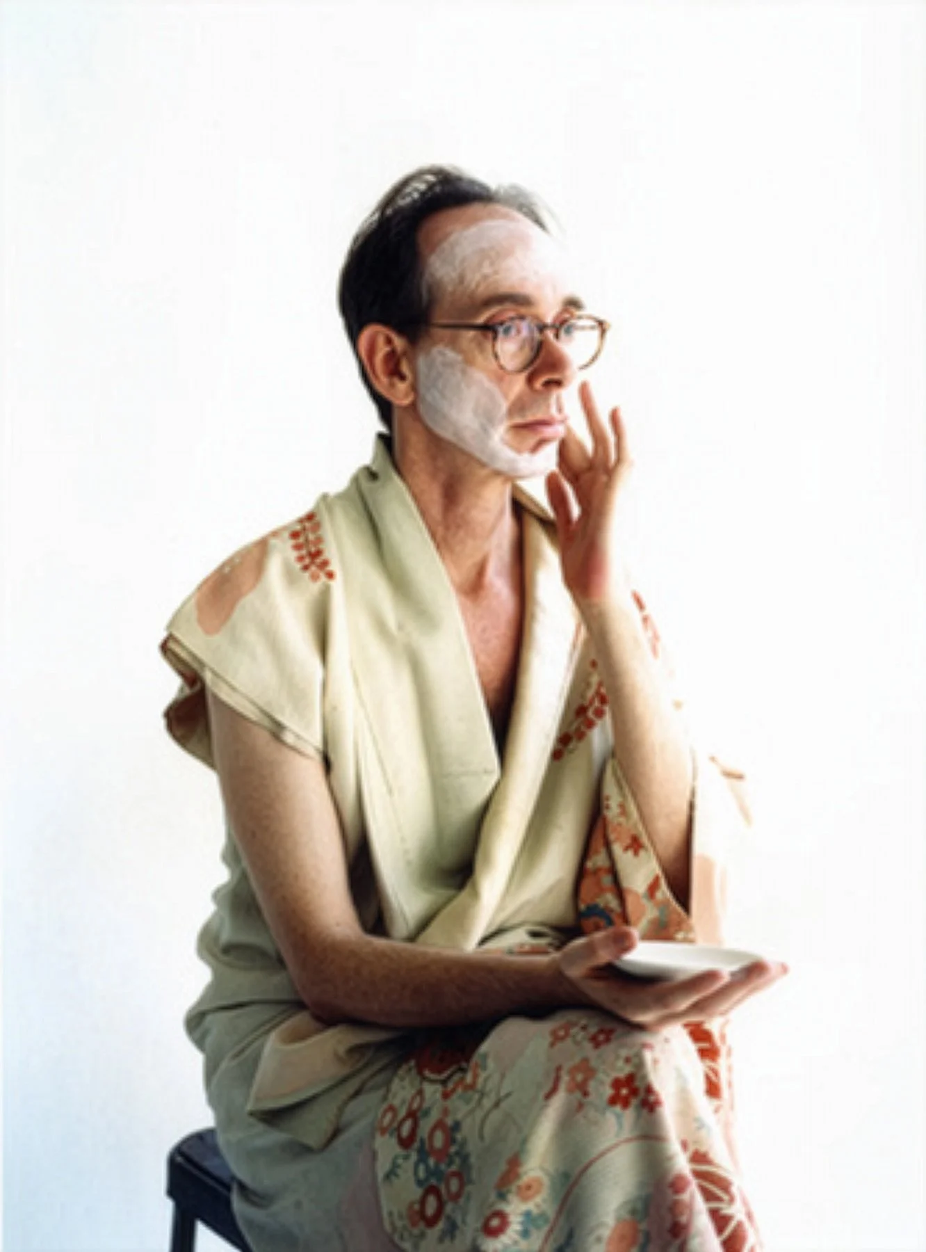 A person with glasses and a bath mask in a beige floral bathrobe sitting on a stool, holding a small plate, with a white background.