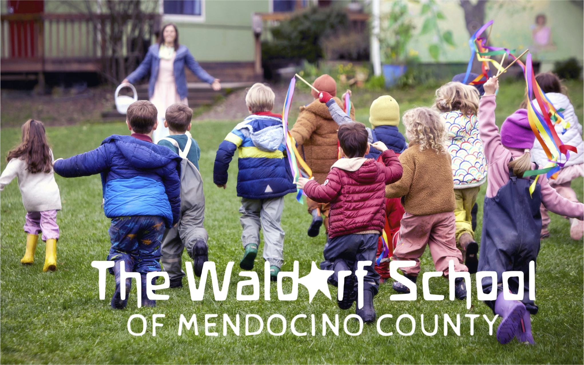 Children running towards a woman, possibly a teacher, outside on a grassy field during daytime. Some children hold colorful ribbons, and all are dressed warmly with jackets and boots. The background shows a building and trees. Overlaid text reads 'The Waldorf School of Mendocino County.'
