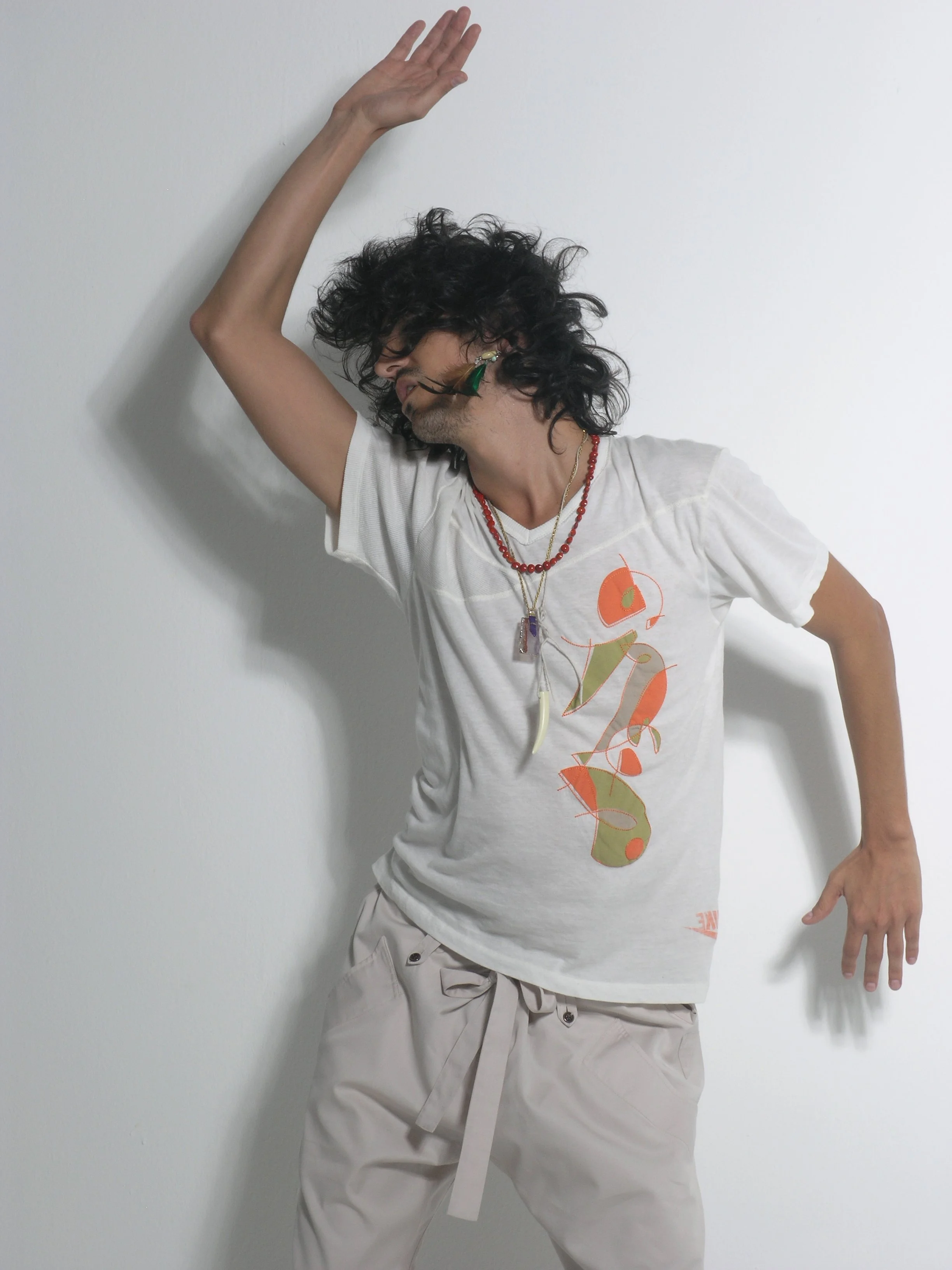 Ricardo Castro, artist with curly hair dancing against a white wall, wearing a graphic Nike t-shirt, beige pants, and layered necklaces. Purple Magazine Concrete Ginga editorial.